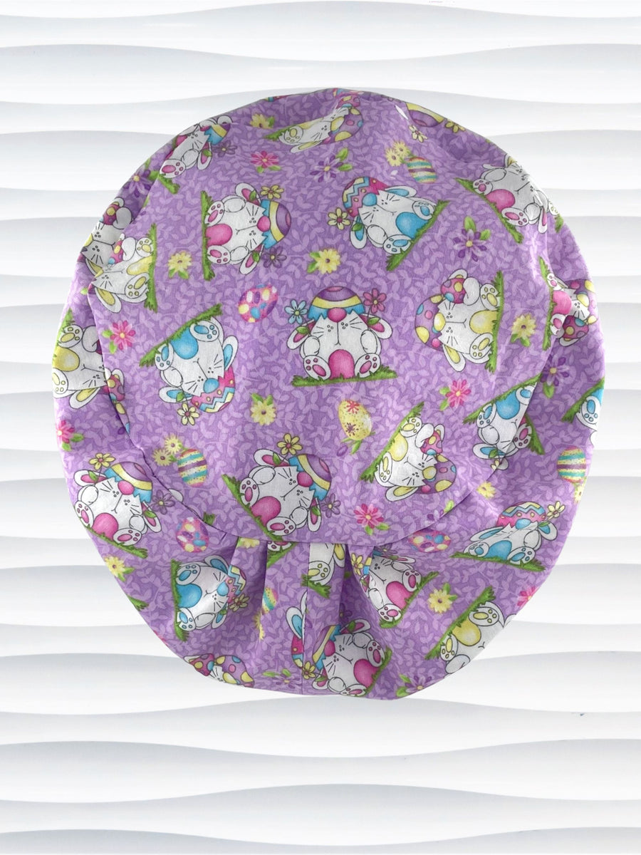 Pixie Euro style surgical scrub cap hat with bunnies wearing cracked egg hats, flowers and easter eggs on light purple lilac cotton fabric.