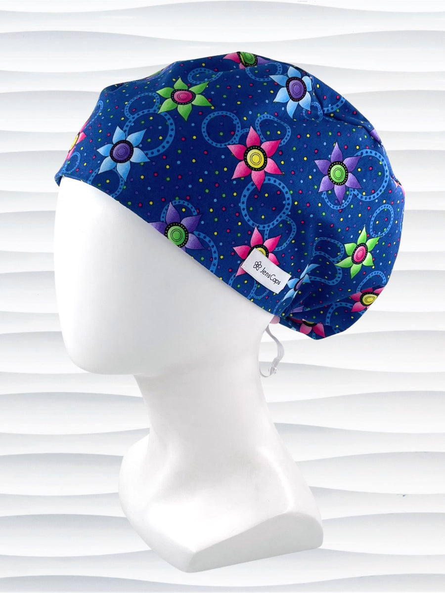 Euro Pixie style surgical scrub cap hat with bright pink, blue, and green flowers and blue circles on dark blue cotton fabric.