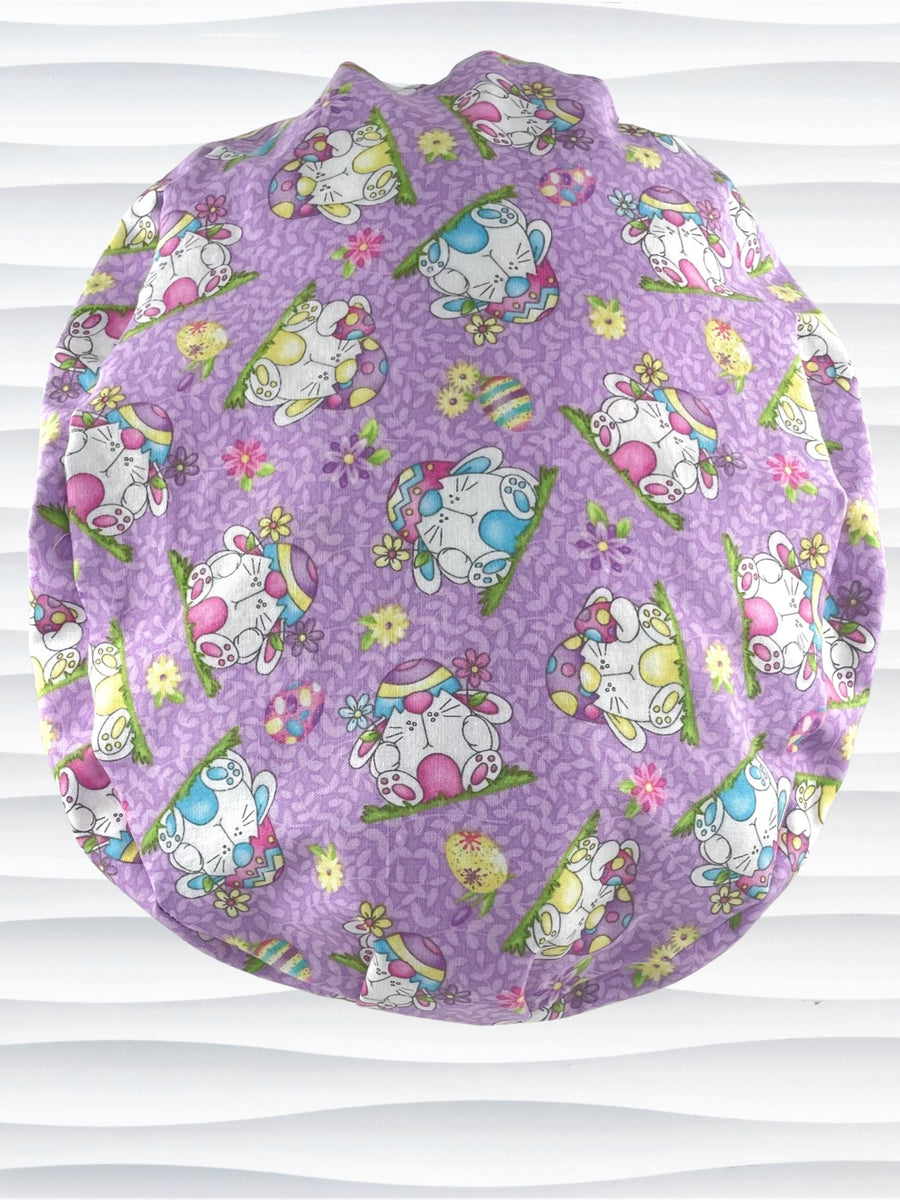 Ponytail style surgical scrub cap hat with bunnies wearing cracked egg hats, flowers and easter eggs on light purple lilac cotton fabric.