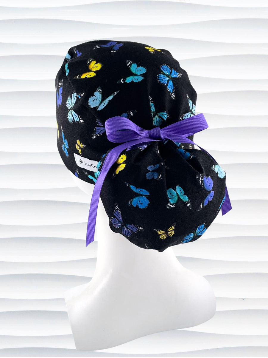Ponytail style surgical scrub cap hat with blue, purple, and yellow butterflies all over black cotton fabric.