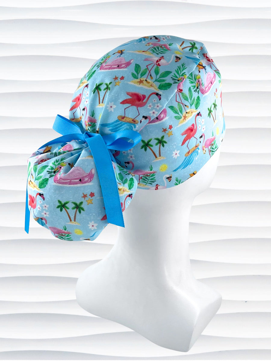 Ponytail surgical scrub cap hat with surfing flamingos, tropical drinks, leis, flowers, and sunglasses on blue cotton fabric.