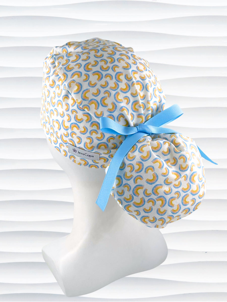 Ponytail style surgical scrub cap hat with an allover print of light blue rainbows on white cotton fabric.