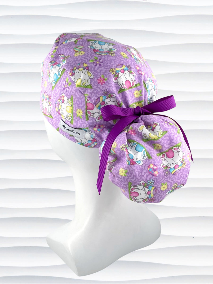 Ponytail style surgical scrub cap hat with bunnies wearing cracked egg hats, flowers and easter eggs on light purple lilac cotton fabric.