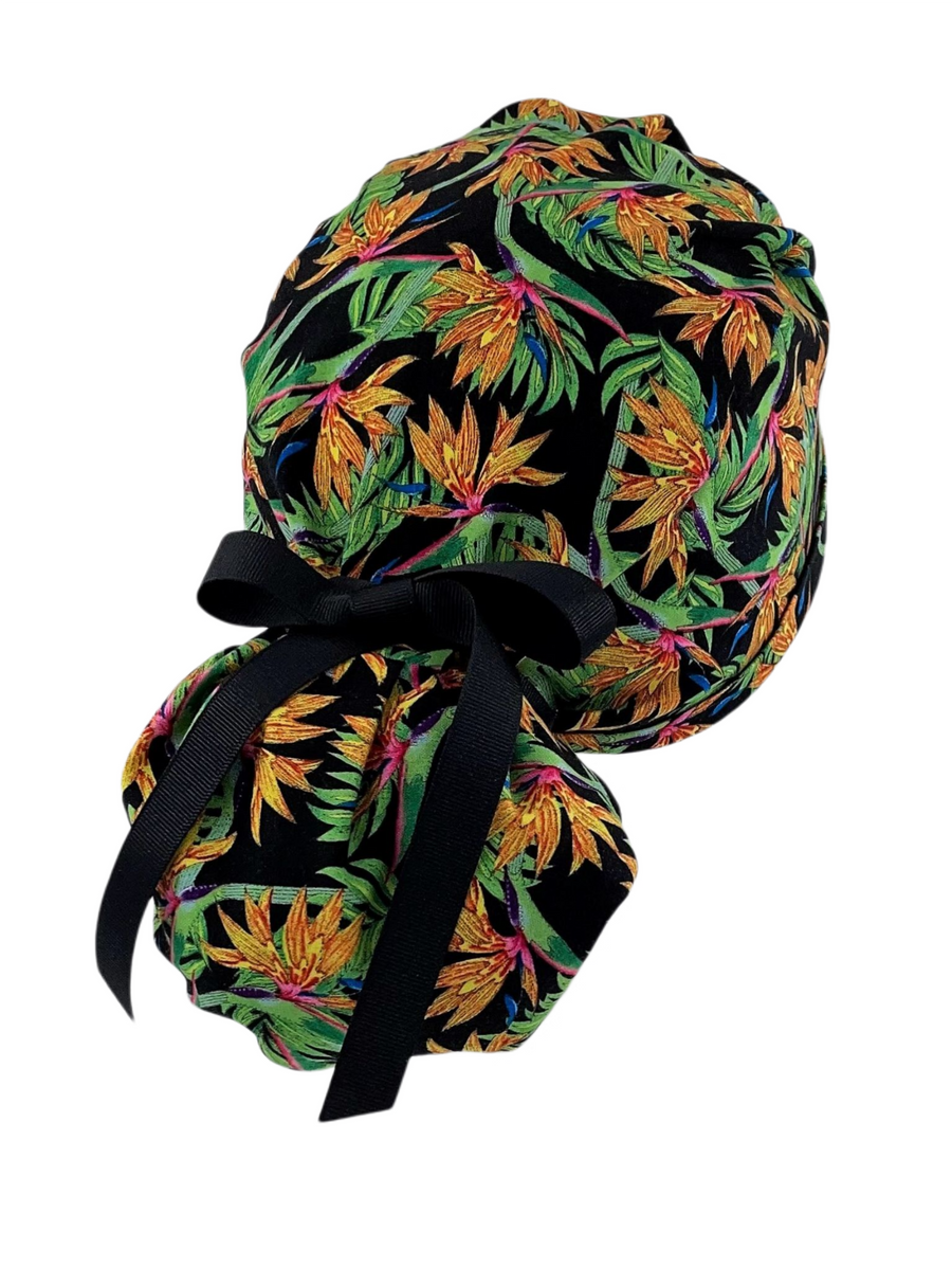 Ponytail style surgical scrub cap hat.  This cotton fabric has an allover print of bird of paradise flowers and stems on a black background and finished with a black or pink grosgrain ribbon
