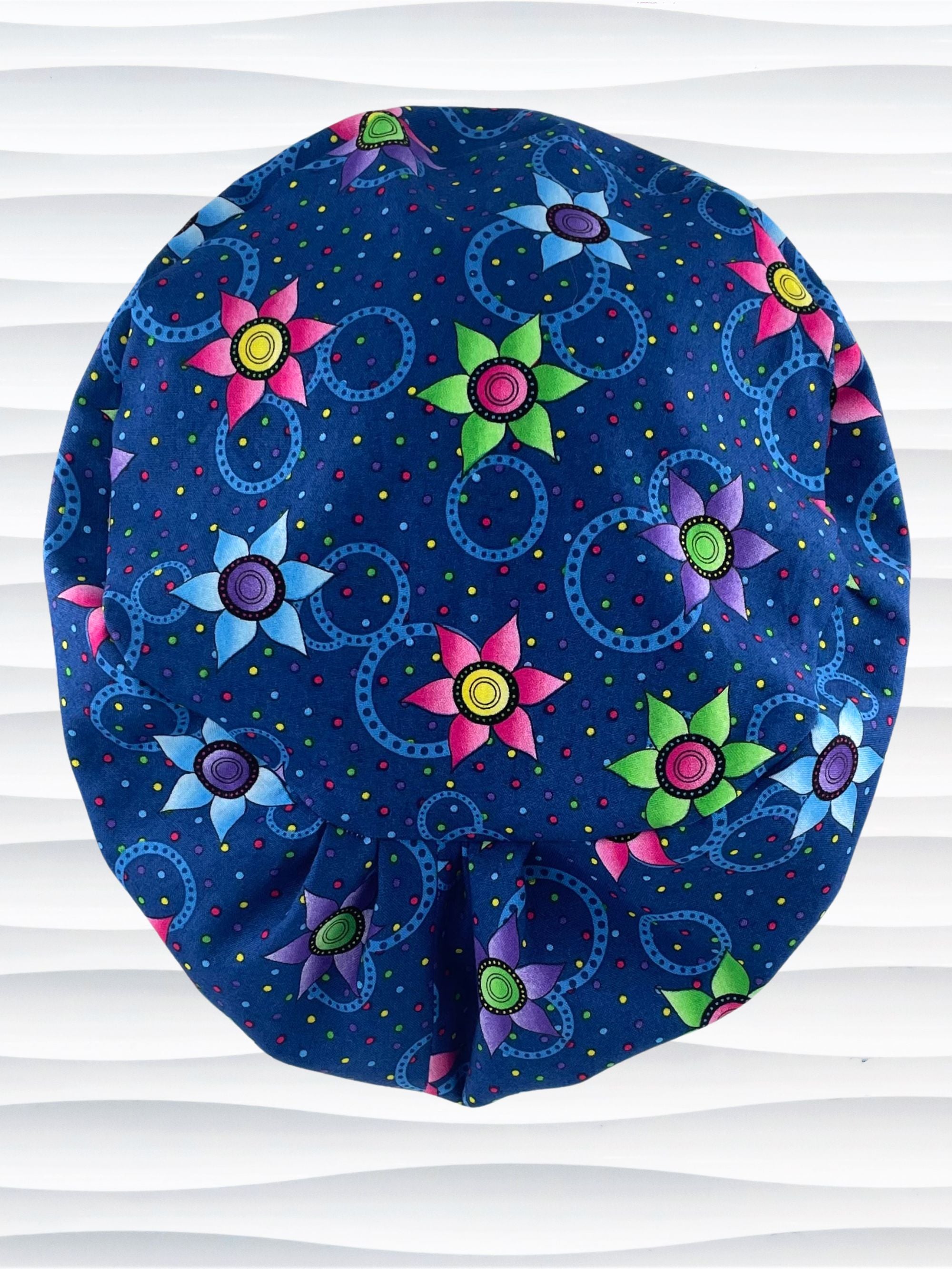 Euro Pixie style surgical scrub cap hat with bright pink, blue, and green flowers and blue circles on dark blue cotton fabric.