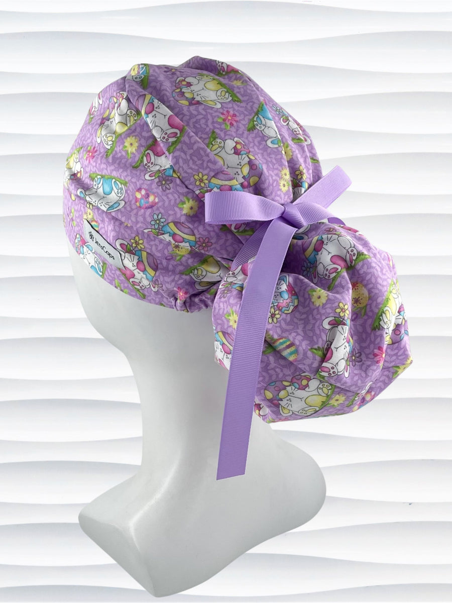 Ponytail style surgical scrub cap hat with bunnies wearing cracked egg hats, flowers and easter eggs on light purple lilac cotton fabric.