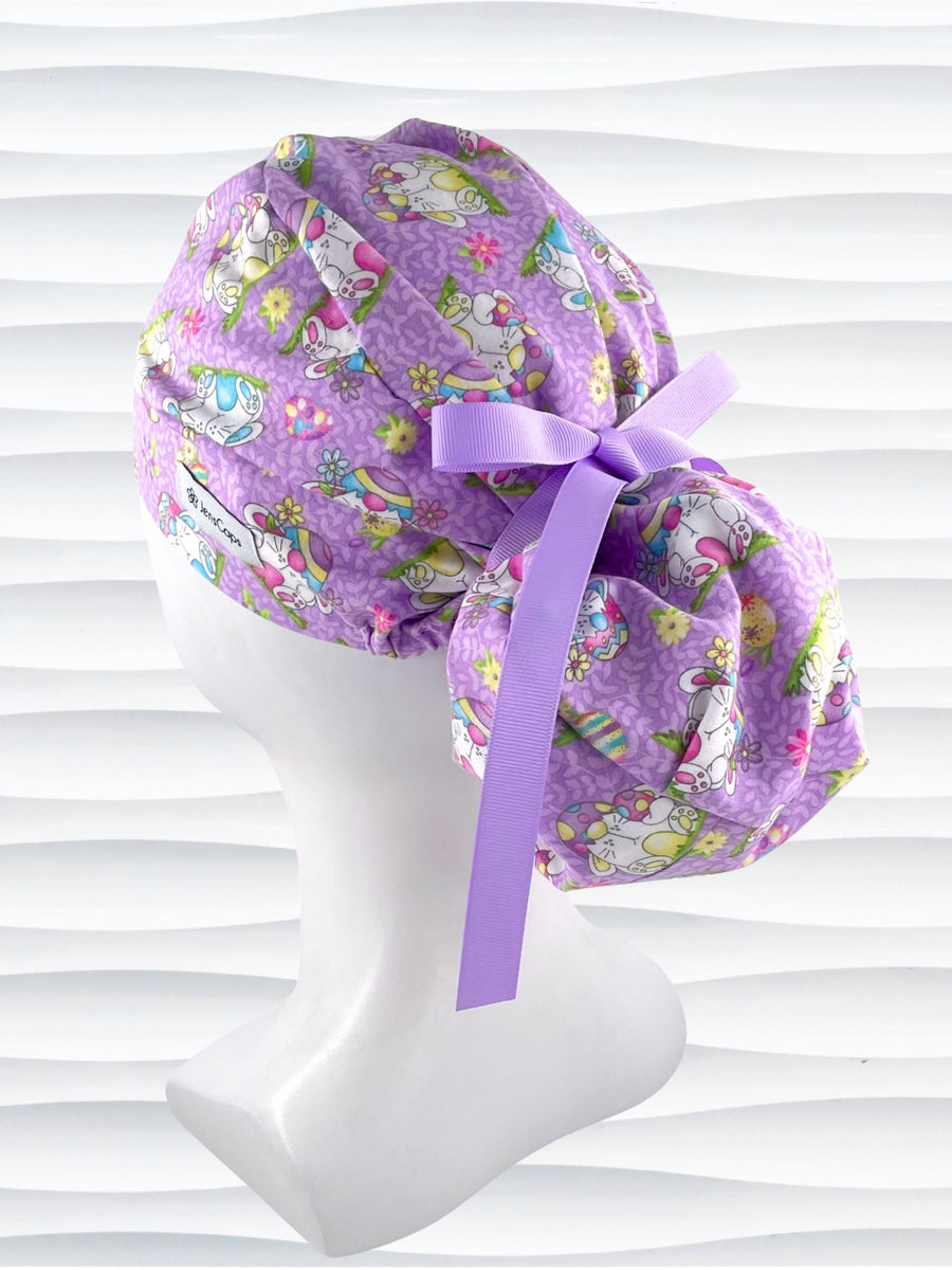 Ponytail style surgical scrub cap hat with bunnies wearing cracked egg hats, flowers and easter eggs on light purple lilac cotton fabric.