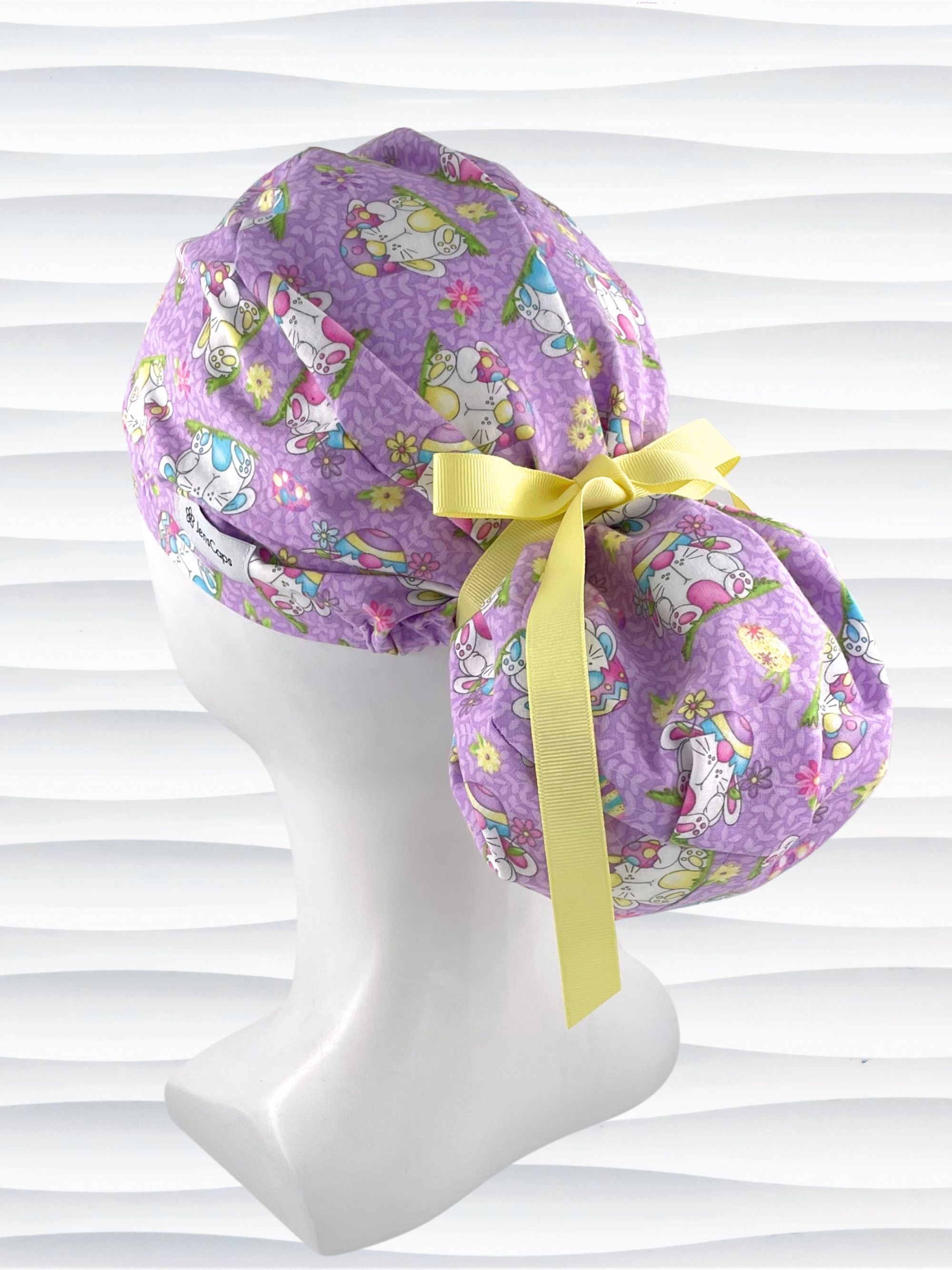 Ponytail style surgical scrub cap hat with bunnies wearing cracked egg hats, flowers and easter eggs on light purple lilac cotton fabric.