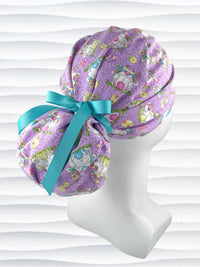 Ponytail style surgical scrub cap hat with bunnies wearing cracked egg hats, flowers and easter eggs on light purple lilac cotton fabric.