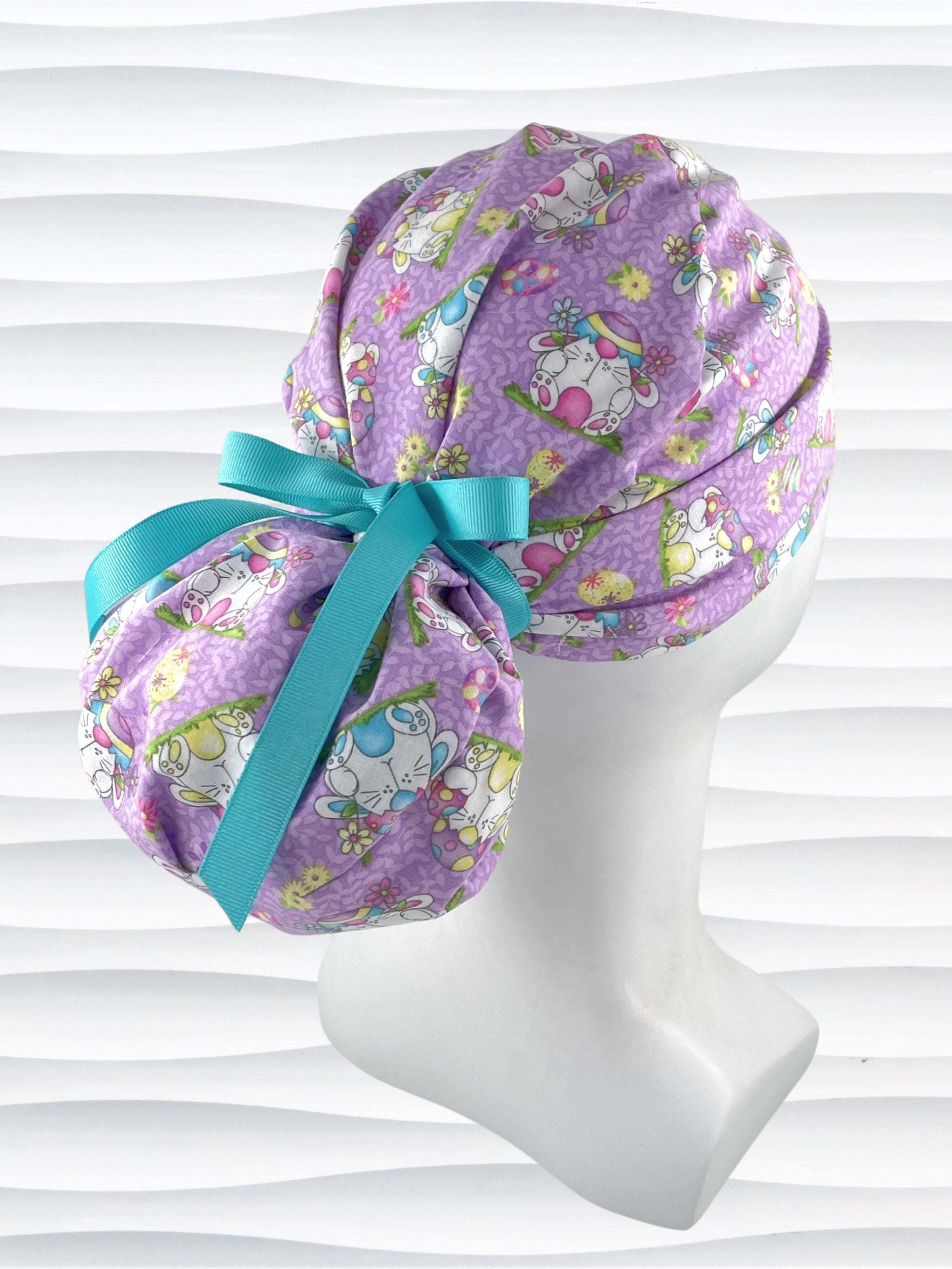 Ponytail style surgical scrub cap hat with bunnies wearing cracked egg hats, flowers and easter eggs on light purple lilac cotton fabric.