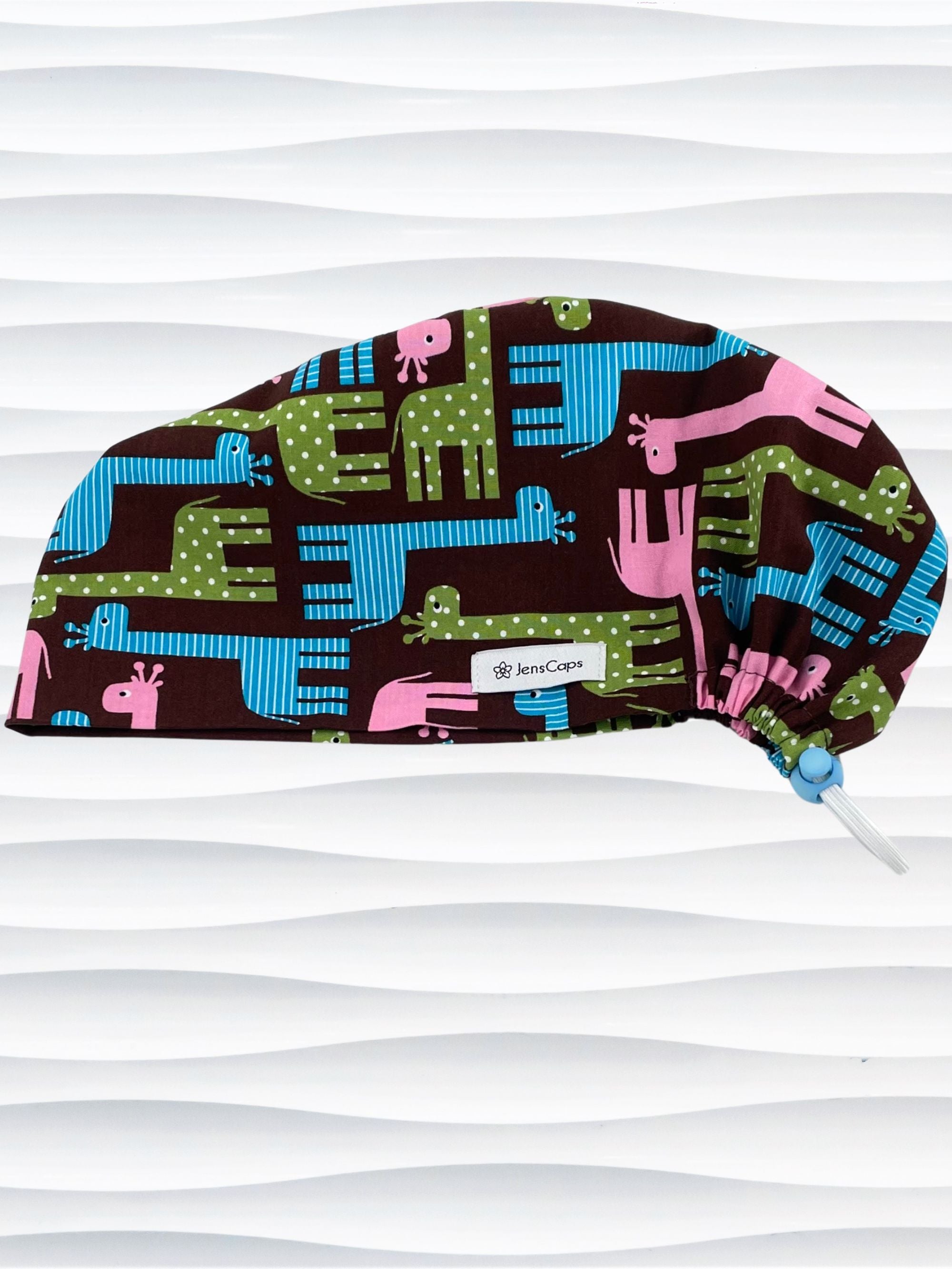 Euro Pixie style surgical scrub cap with cute giraffes with stripes or dots in blue, pink, and green all over a dark brown background on cotton fabric.