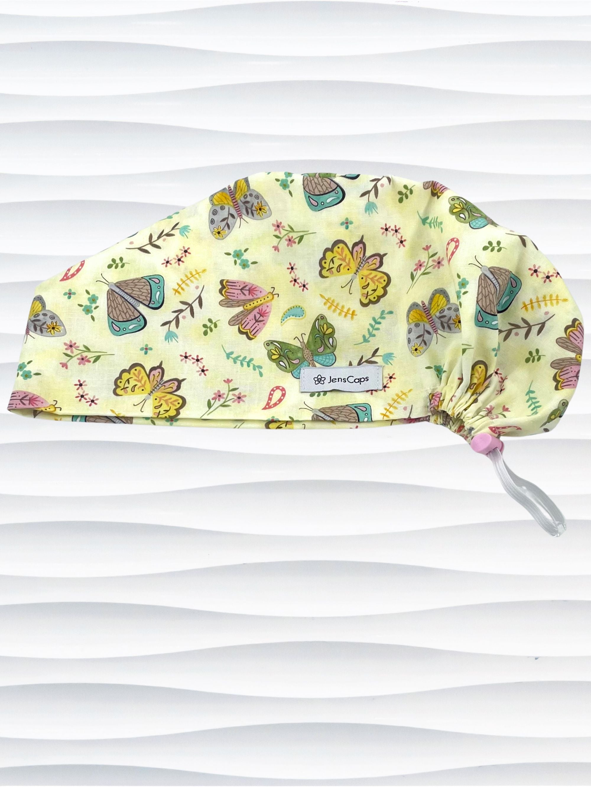 Euro Pixie style surgical scrub cap with butterflies, moths, and flowers in pink, blue, green, and yellow all over a light yellow cotton fabric.