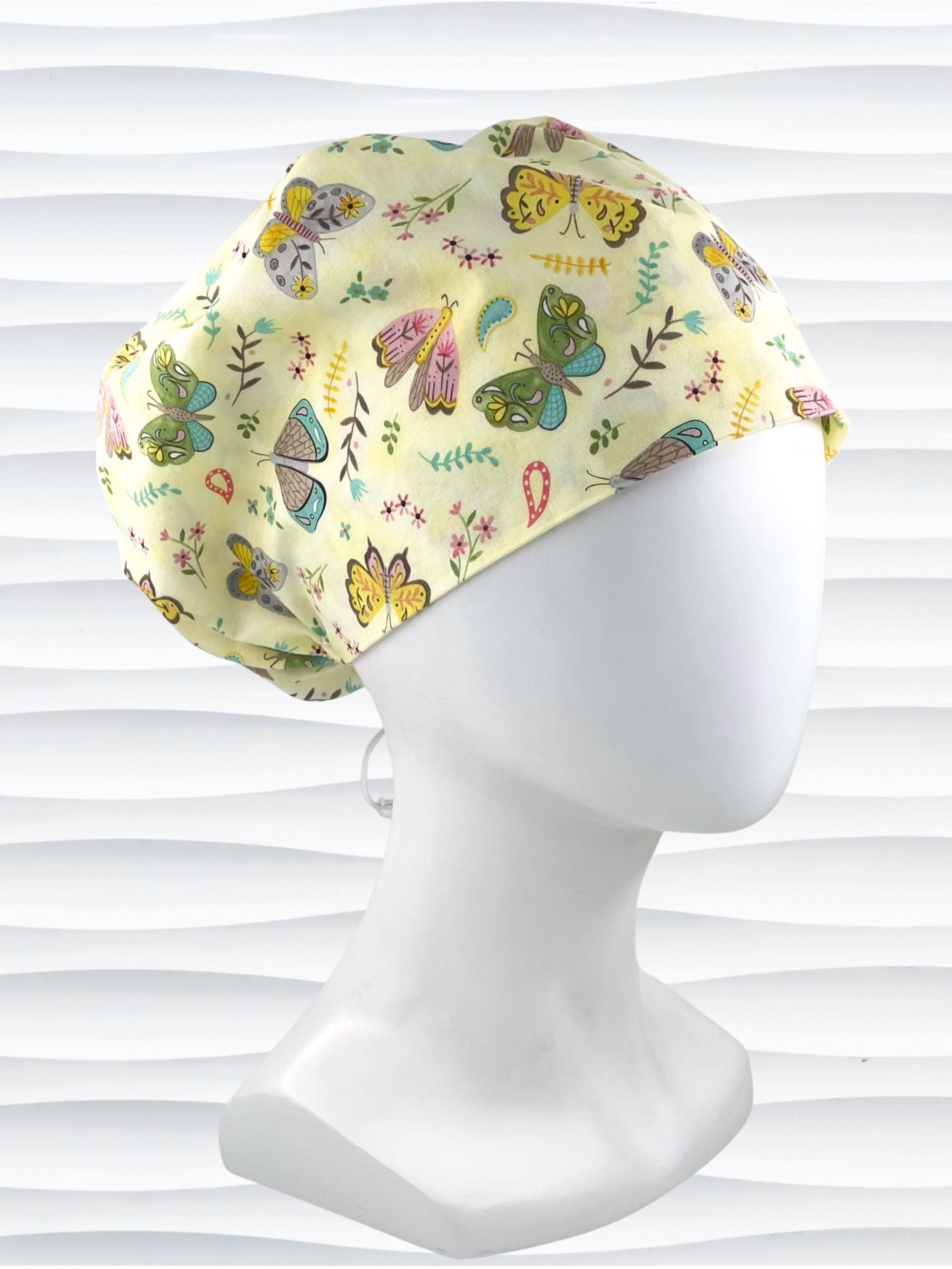 Euro Pixie style surgical scrub cap with butterflies, moths, and flowers in pink, blue, green, and yellow all over a light yellow cotton fabric.