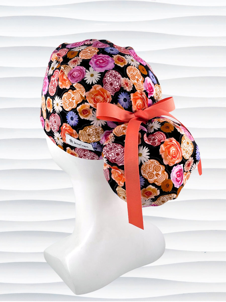 Ponytail style surgical scrub cap hat with sugar skulls and flowers in pink, purple, red, orange, and white on black background cotton fabric.