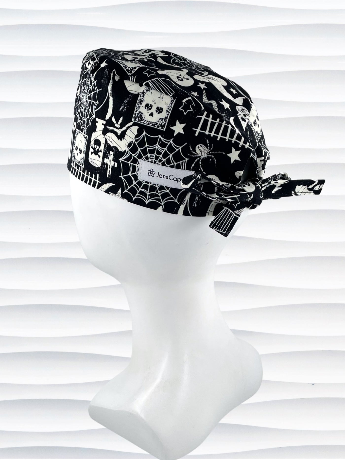 A black and white Halloween-themed tie back style scrub cap with a spiderweb pattern, skulls, and ghosts, displayed on a white mannequin head. The brand name 'JensCaps' is visible on a white label sewn onto the side of the cap.