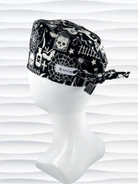 A black and white Halloween-themed tie back style scrub cap with a spiderweb pattern, skulls, and ghosts, displayed on a white mannequin head. The brand name 'JensCaps' is visible on a white label sewn onto the side of the cap.
