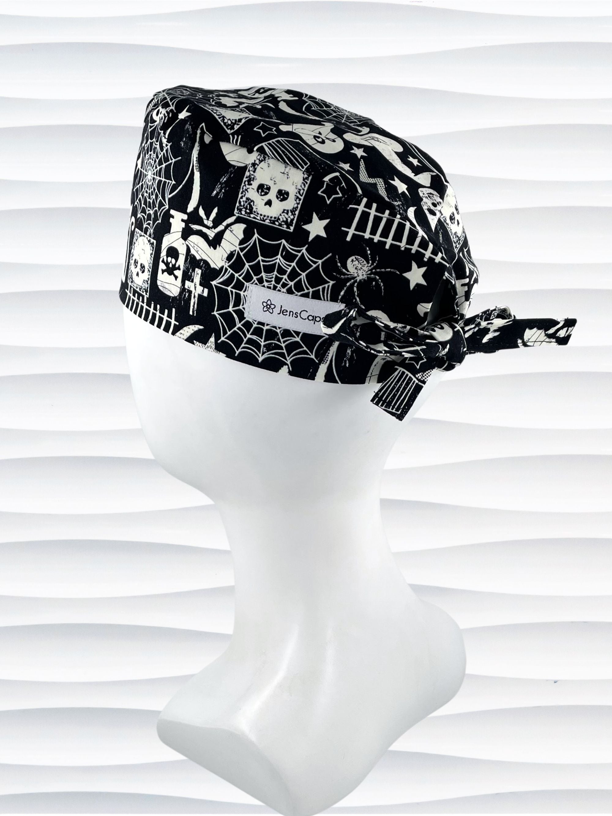 A black and white Halloween-themed tie back style scrub cap with a spiderweb pattern, skulls, and ghosts, displayed on a white mannequin head. The brand name 'JensCaps' is visible on a white label sewn onto the side of the cap.