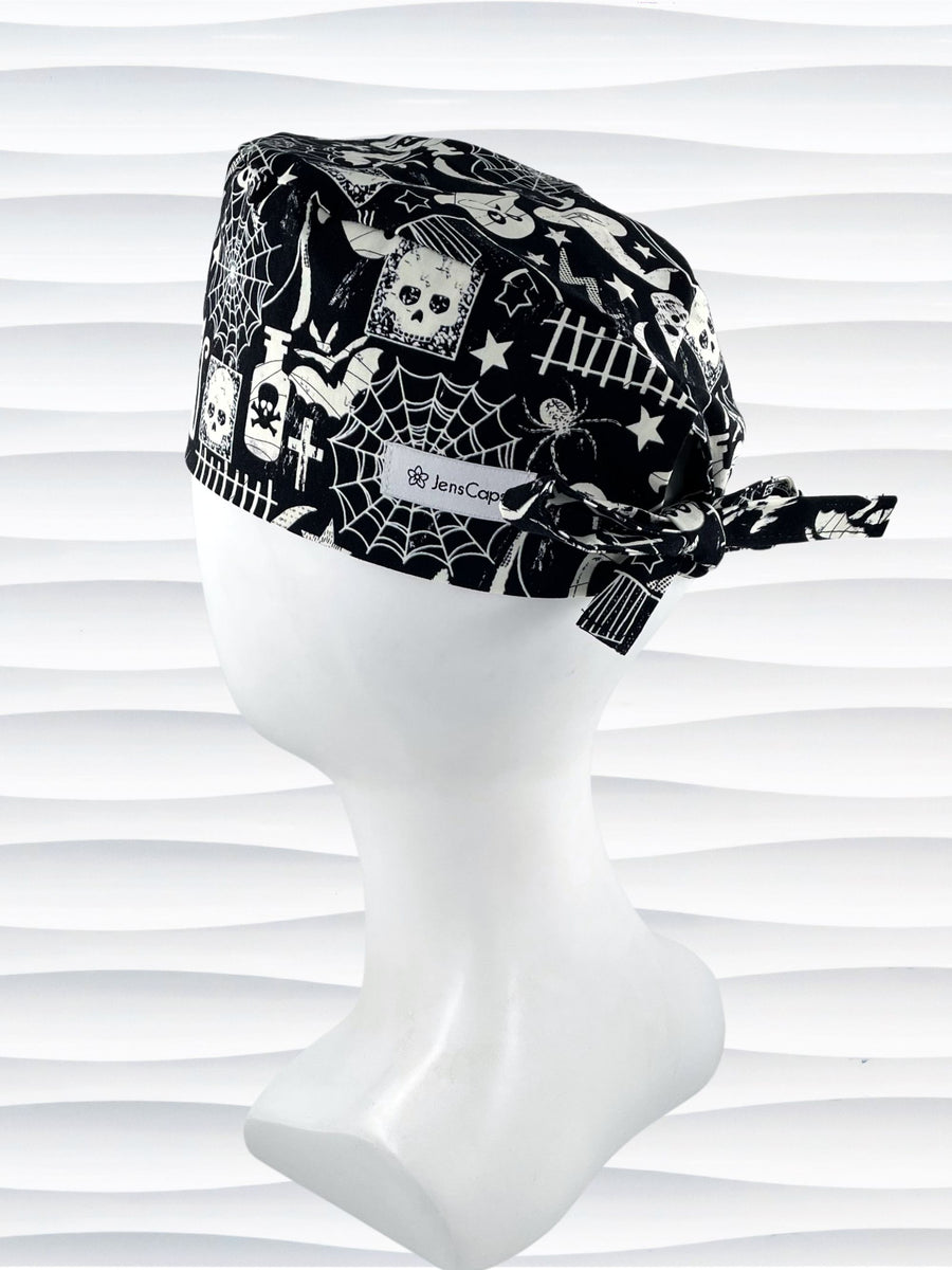 A black and white Halloween-themed tie back style scrub cap with a spiderweb pattern, skulls, and ghosts, displayed on a white mannequin head. The brand name 'JensCaps' is visible on a white label sewn onto the side of the cap.