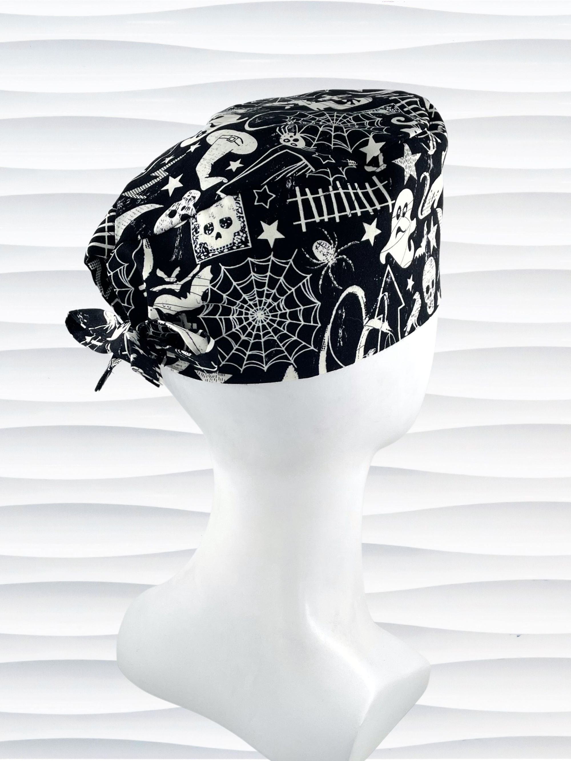 Black and white Halloween-themed Tie back surgical scrub cap with a spiderweb, skull, and ghost pattern, displayed on a white mannequin head