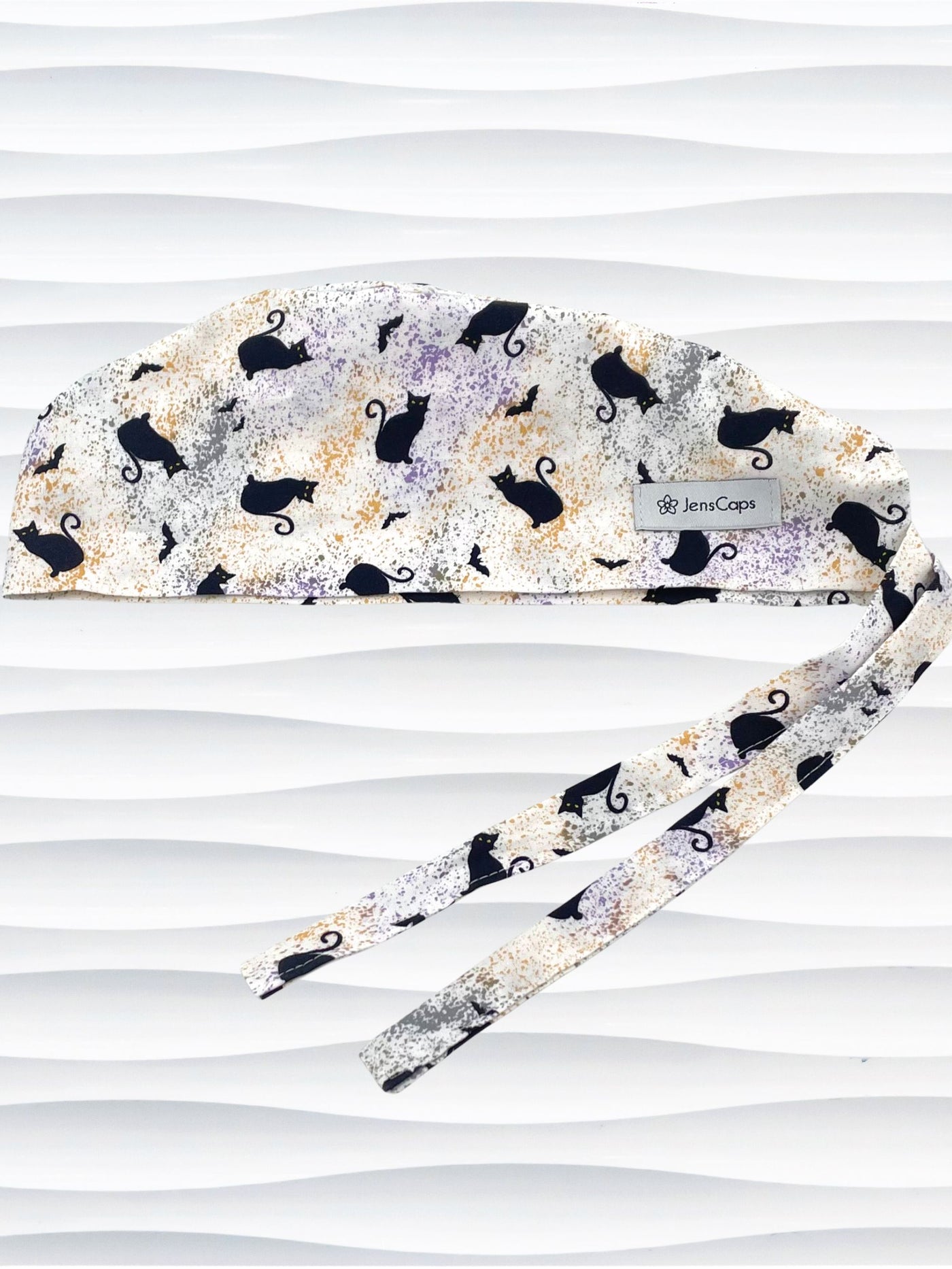 A tie-back style surgical scrub cap by JensCaps with a black cat silhouette pattern and subtle purple and gold speckles, displayed on a white mannequin head against a wavy white background