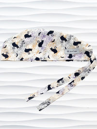 A tie-back style surgical scrub cap by JensCaps with a black cat silhouette pattern and subtle purple and gold speckles, displayed on a white mannequin head against a wavy white background