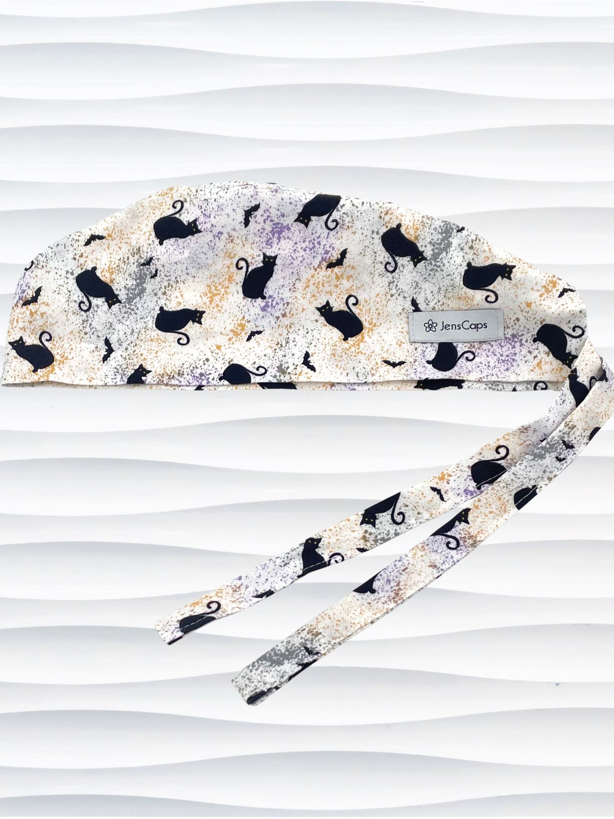 A tie-back style surgical scrub cap by JensCaps with a black cat silhouette pattern and subtle purple and gold speckles, displayed on a white mannequin head against a wavy white background