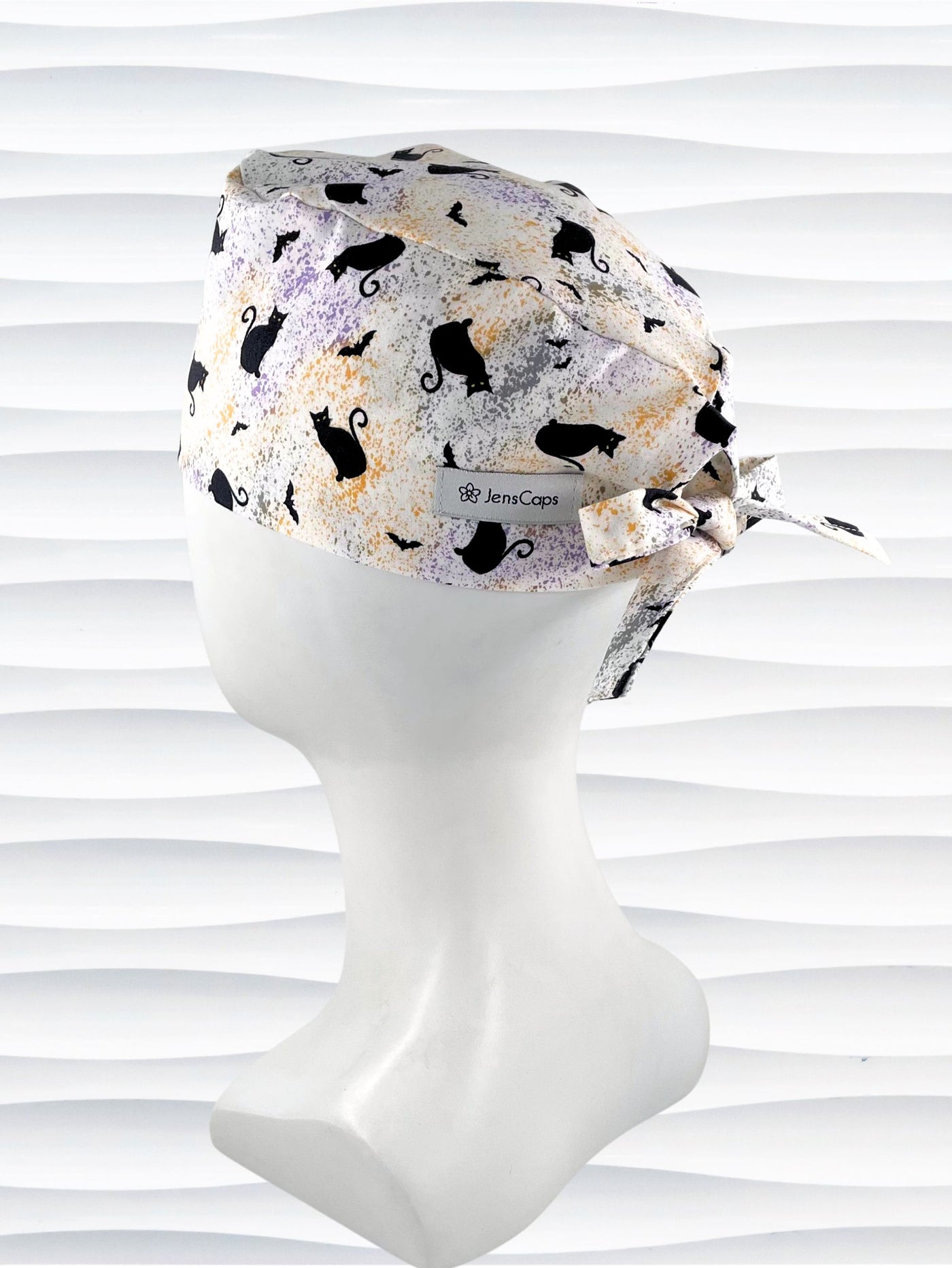 A tie-back style surgical scrub cap by JensCaps with a black cat silhouette pattern and subtle purple and gold speckles, displayed on a white mannequin head against a wavy white background