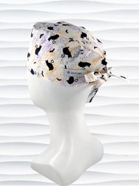 A tie-back style surgical scrub cap by JensCaps with a black cat silhouette pattern and subtle purple and gold speckles, displayed on a white mannequin head against a wavy white background