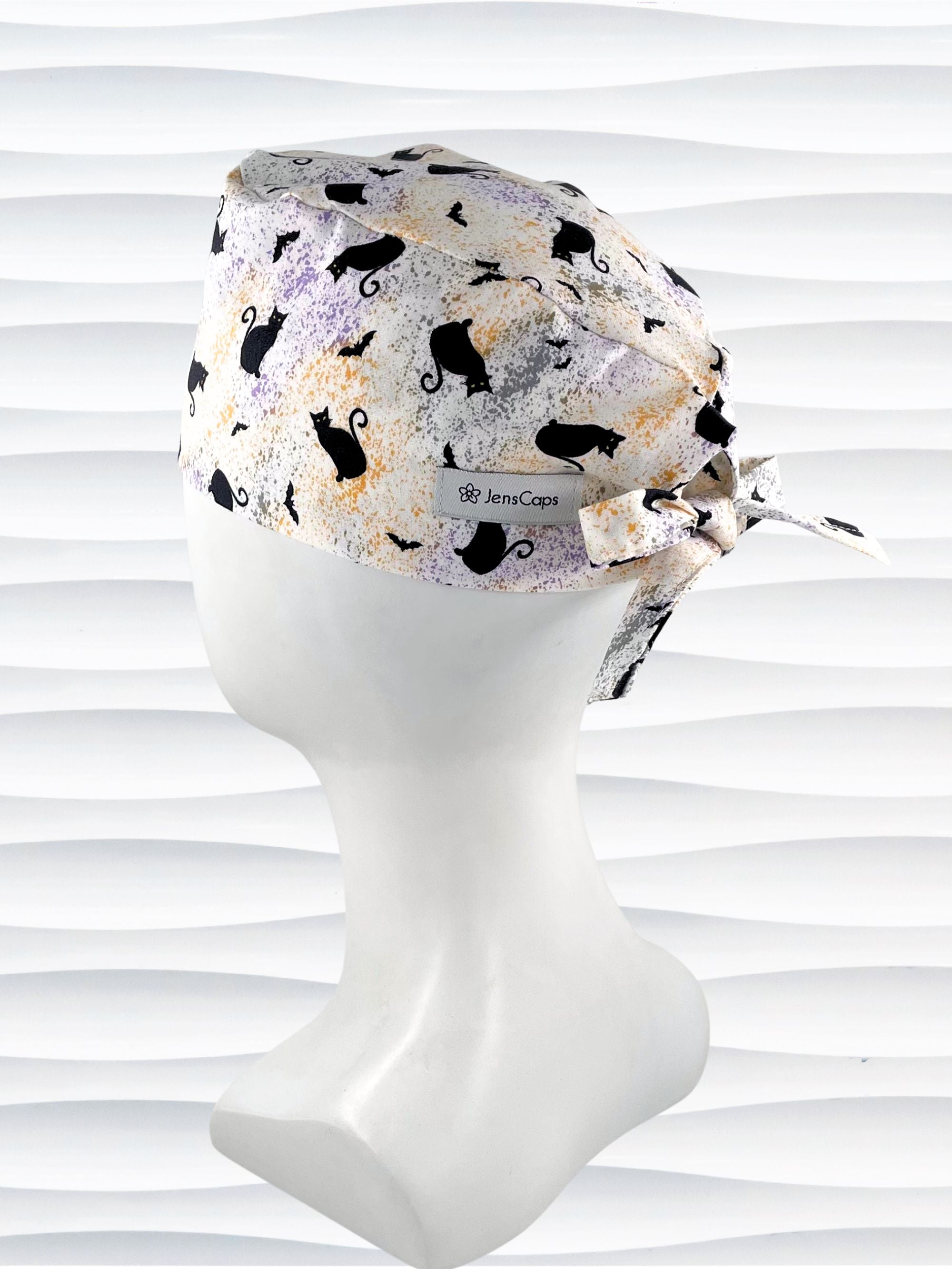 A tie-back style surgical scrub cap by JensCaps with a black cat silhouette pattern and subtle purple and gold speckles, displayed on a white mannequin head against a wavy white background