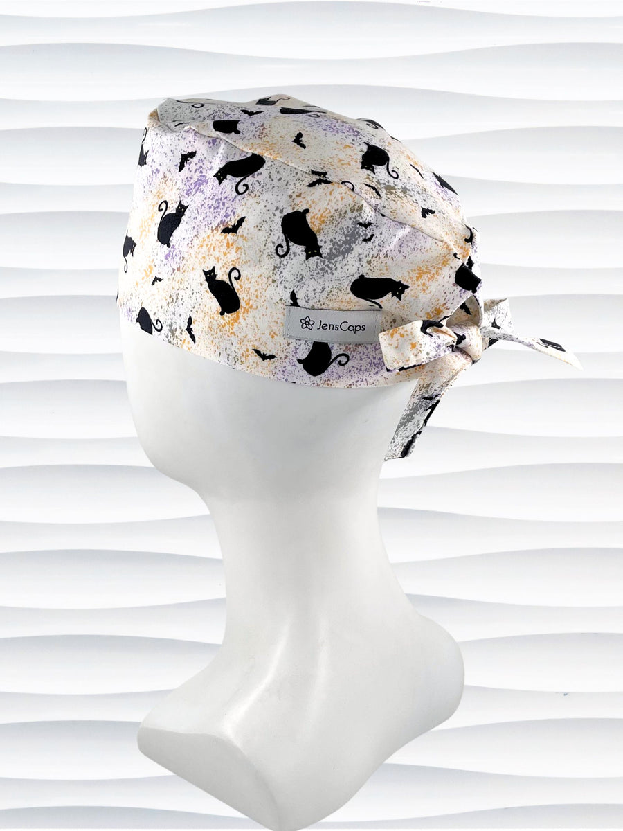 A tie-back style surgical scrub cap by JensCaps with a black cat silhouette pattern and subtle purple and gold speckles, displayed on a white mannequin head against a wavy white background