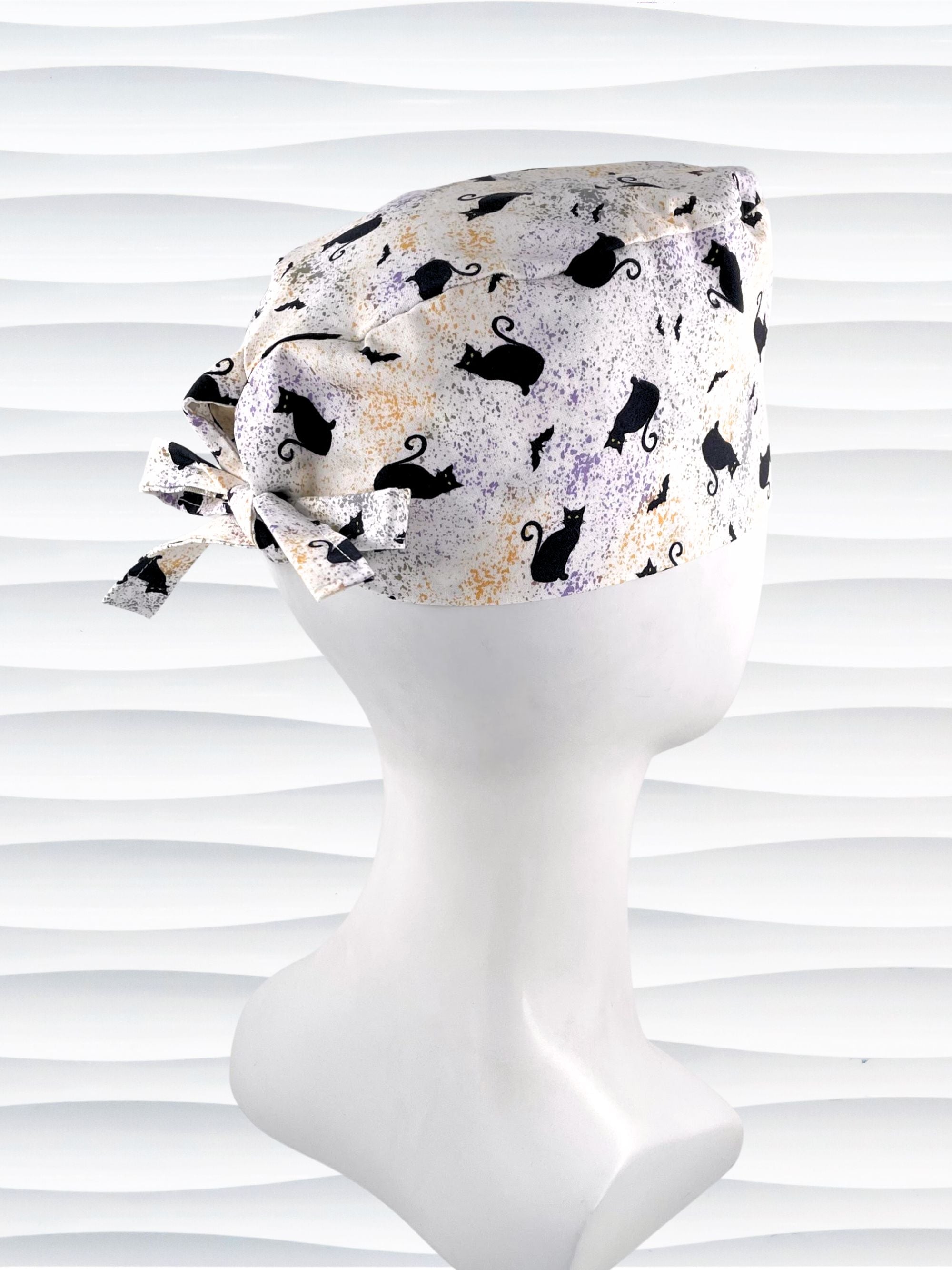 A tie-back style surgical scrub cap by JensCaps with a black cat silhouette pattern and subtle purple and gold speckles, displayed on a white mannequin head against a wavy white background