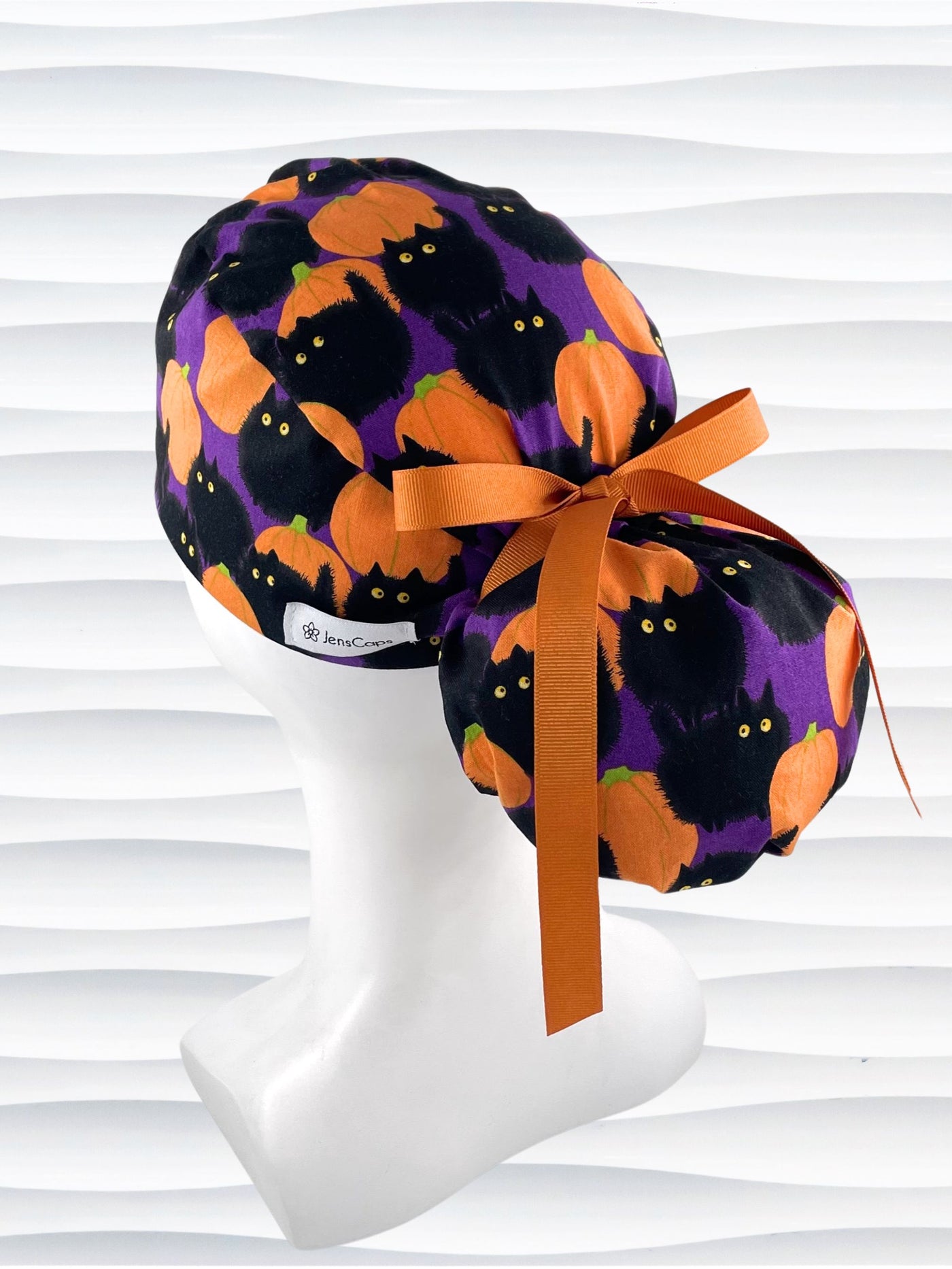 Ponytail style surgical scrub cap hat with puffy black cats and orange pumpkins all over this purple background cotton fabric.