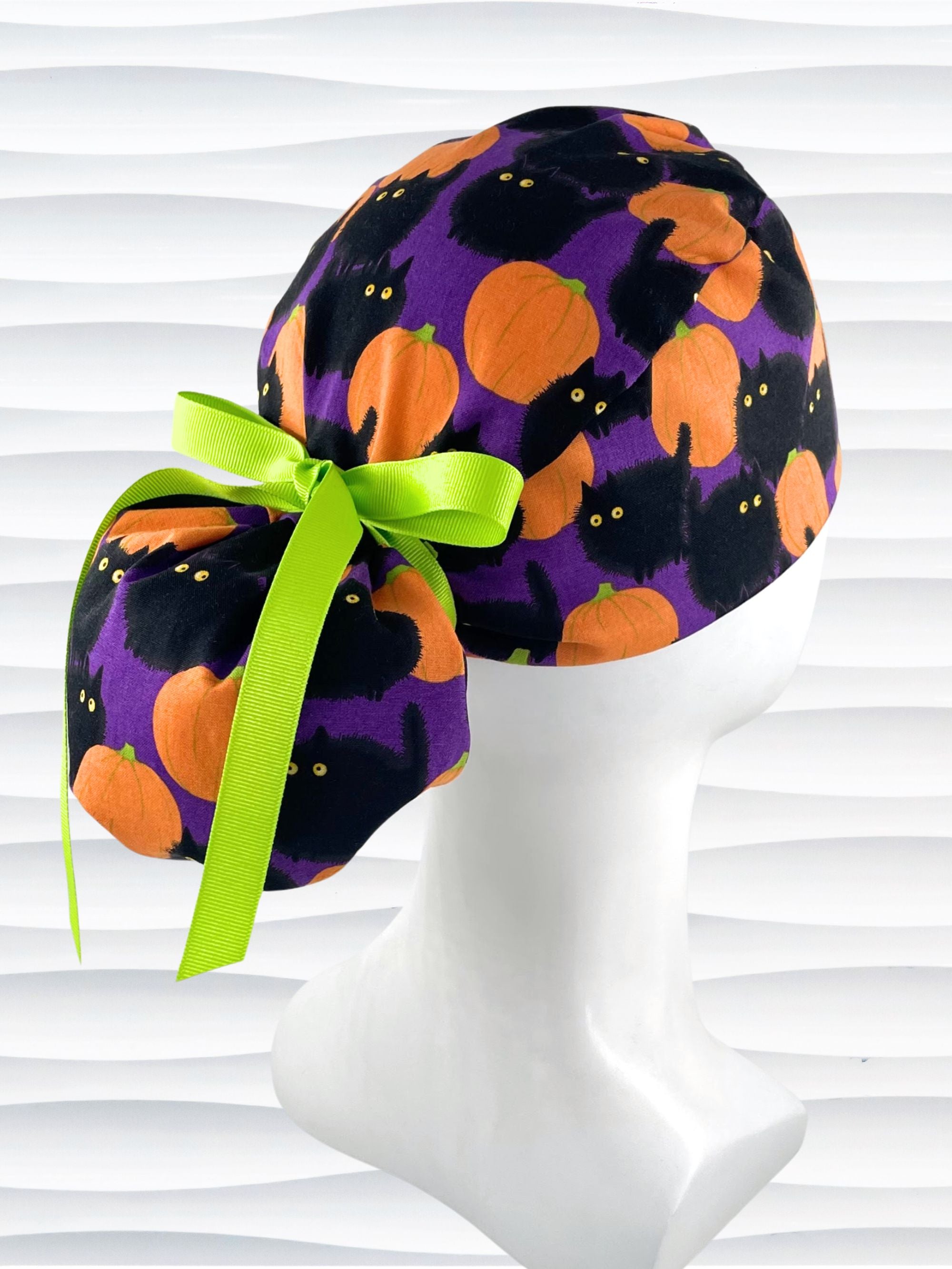 Ponytail style surgical scrub cap hat with puffy black cats and orange pumpkins all over this purple background cotton fabric.