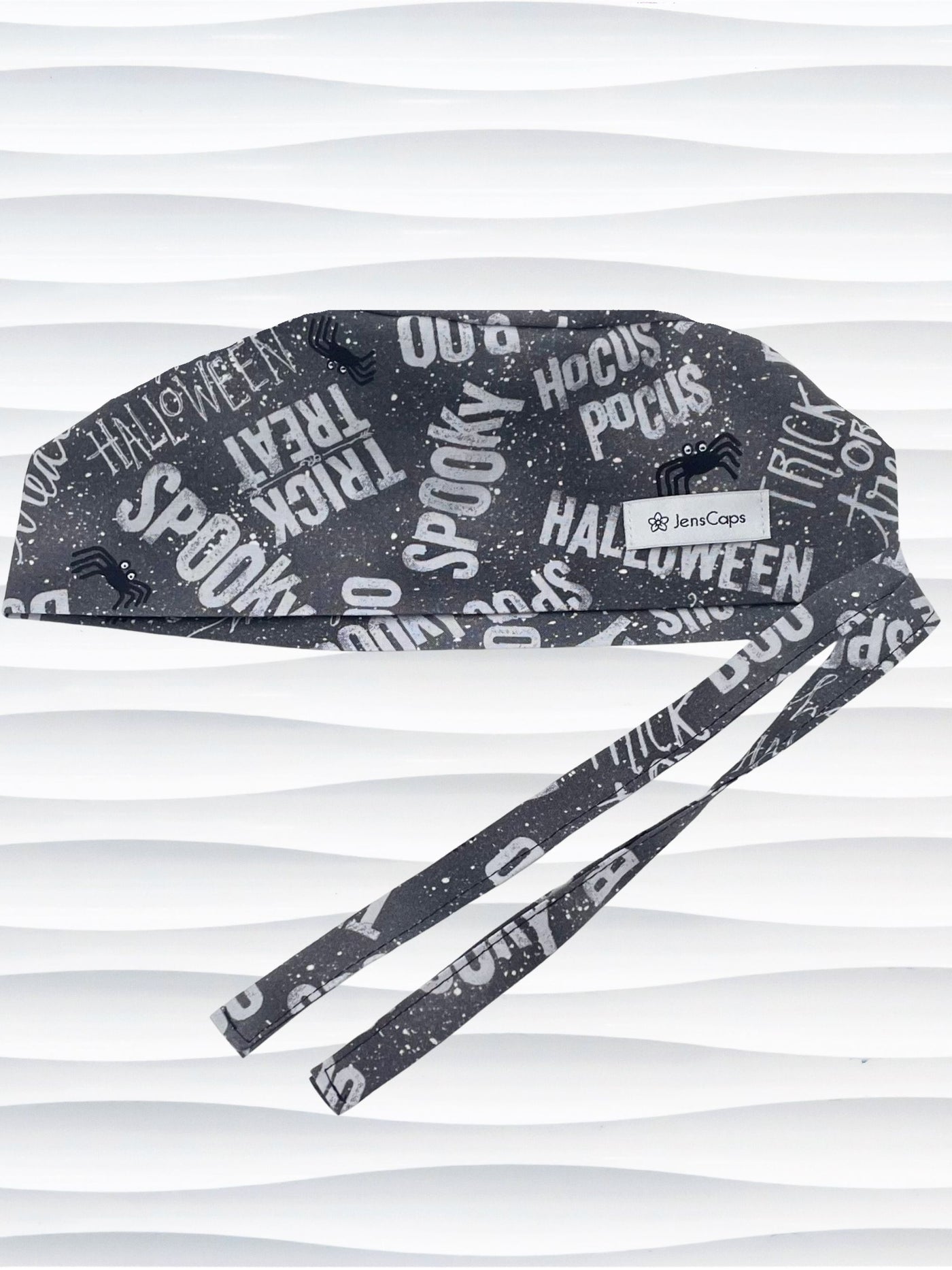 A dark gray Halloween-themed tie-back style scrub cap by JensCaps with white text patterns spelling 'Spooky', 'Halloween', 'Trick or Treat', and 'Pocus', featuring adjustable ties 