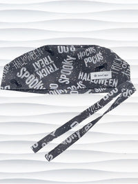 A dark gray Halloween-themed tie-back style scrub cap by JensCaps with white text patterns spelling 'Spooky', 'Halloween', 'Trick or Treat', and 'Pocus', featuring adjustable ties 