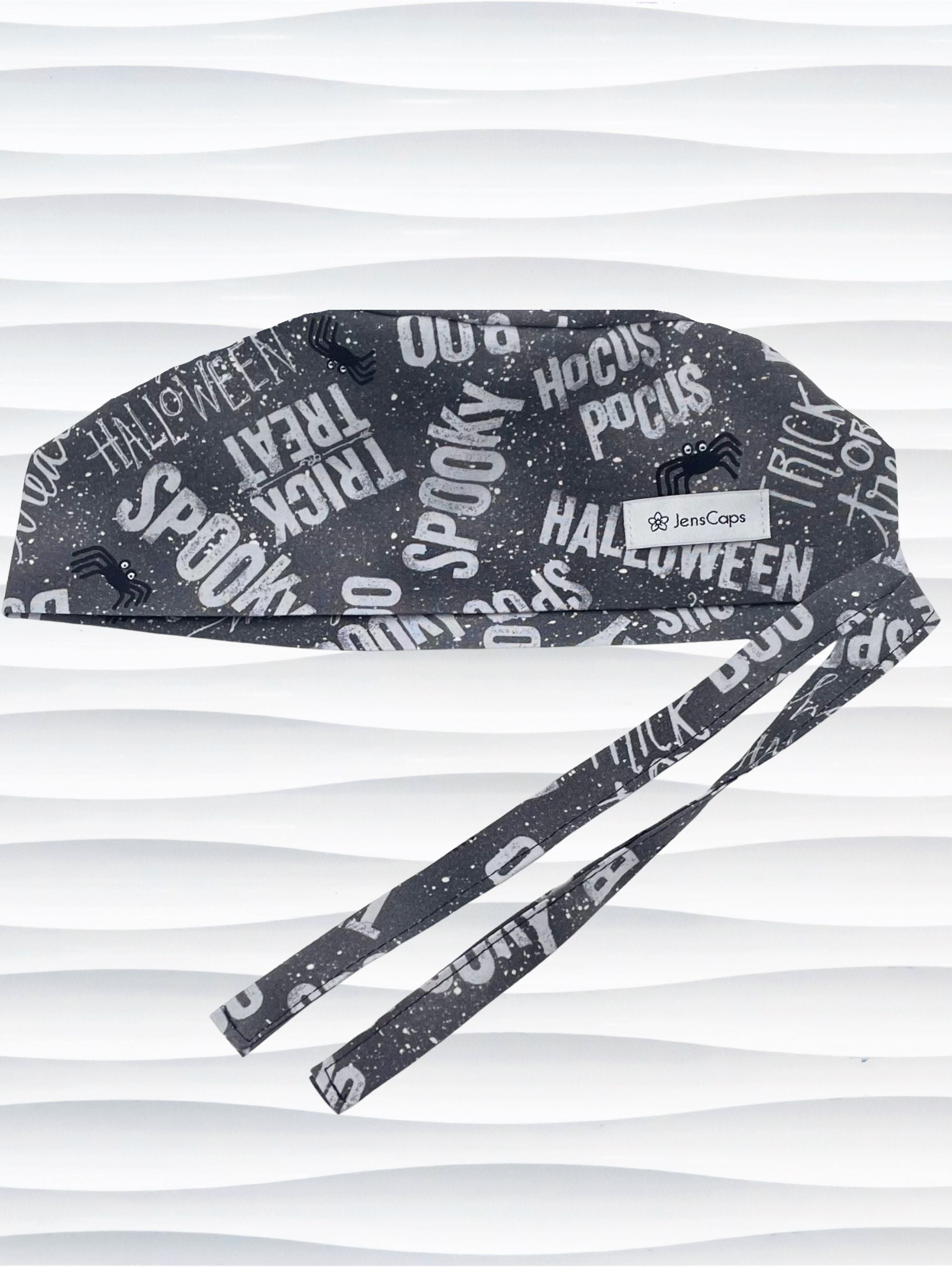 A dark gray Halloween-themed tie-back style scrub cap by JensCaps with white text patterns spelling 'Spooky', 'Halloween', 'Trick or Treat', and 'Pocus', featuring adjustable ties 