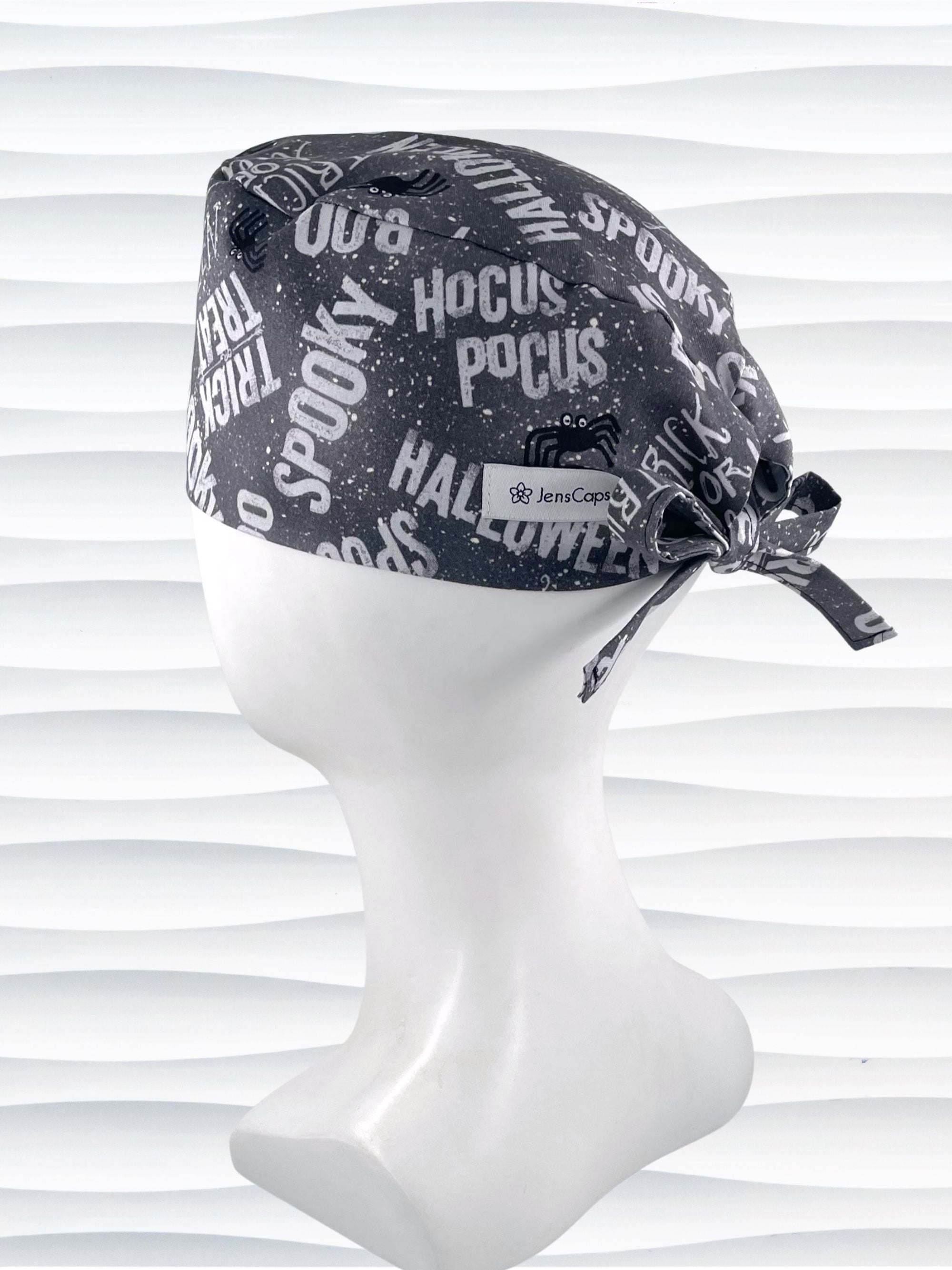 A black and white patterned tie-back style surgical scrub cap by JensCaps with Halloween-themed text like 'Hocus Pocus' and 'Spooky' displayed on a white mannequin head. 