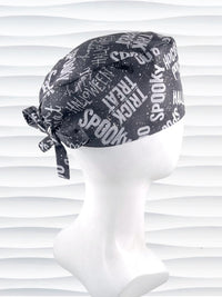 A dark gray Halloween-themed tie-back style scrub cap by JensCaps with white text patterns spelling 'Spooky', 'Halloween', 'Trick or Treat', and 'Pocus', featuring adjustable ties 