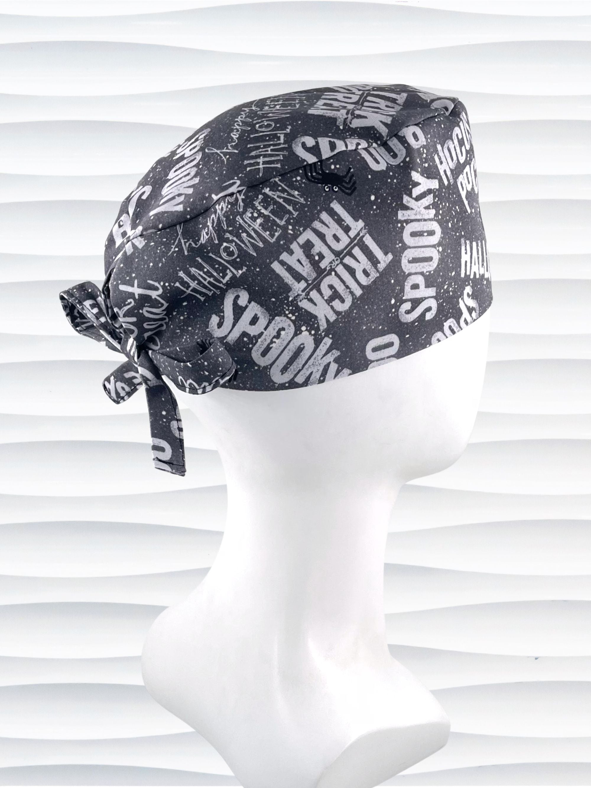 A dark gray Halloween-themed tie-back style scrub cap by JensCaps with white text patterns spelling 'Spooky', 'Halloween', 'Trick or Treat', and 'Pocus', featuring adjustable ties 