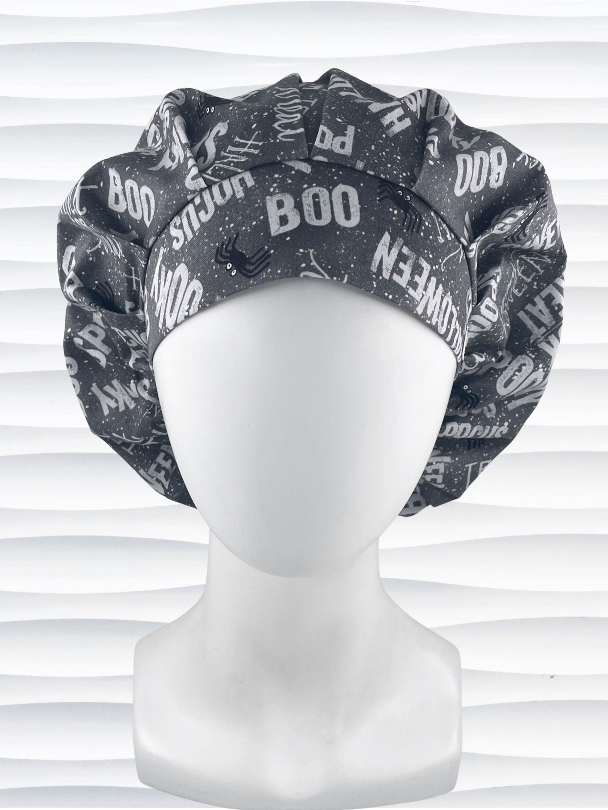 Bouffant style surgical scrub cap by JensCaps with halloween words in white and light gray and black spiders all over gray cotton fabric and displayed on a mannequin head.