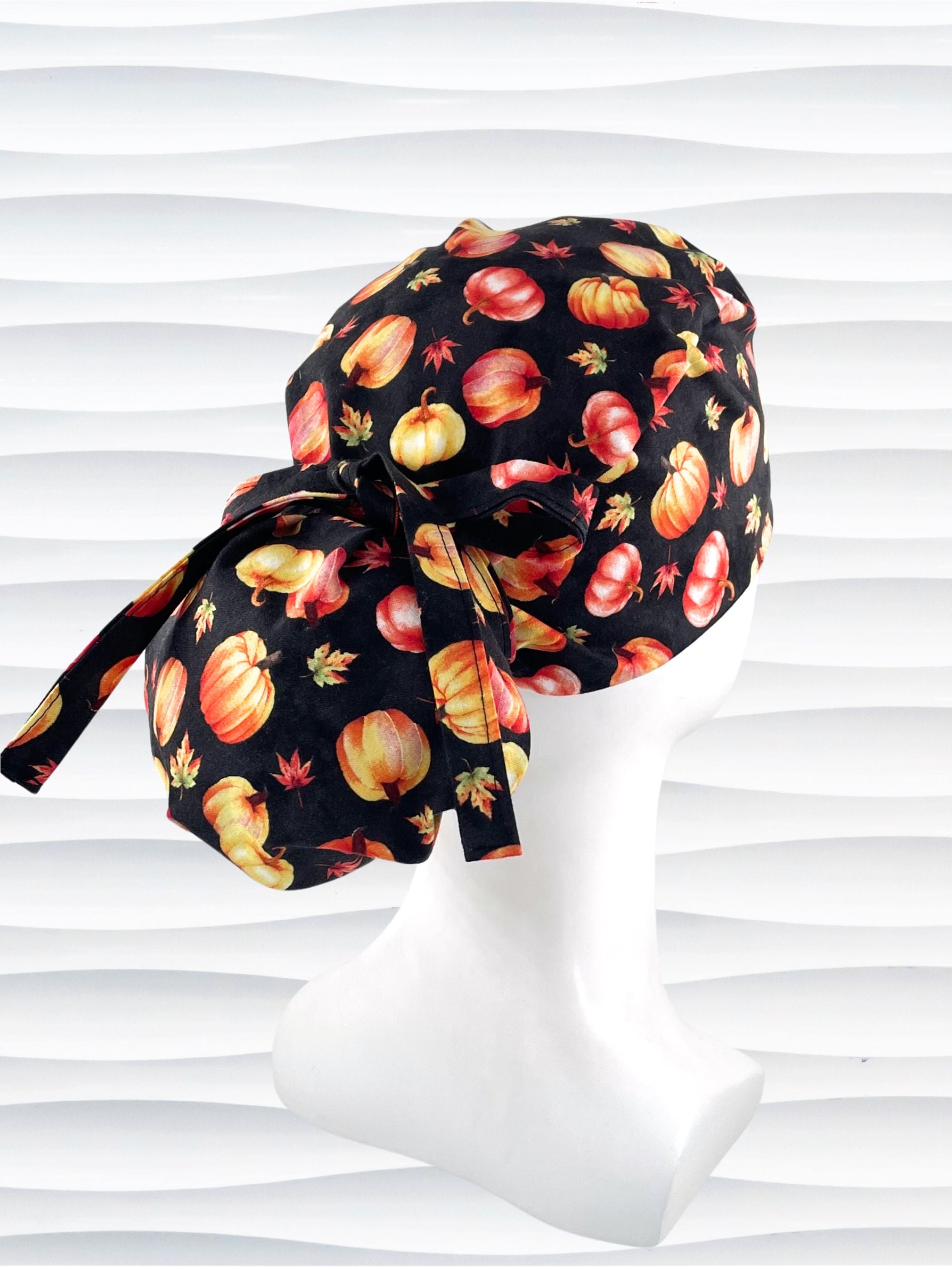 Ponytail style surgical scrub cap hat with orange and yellow pumpkins and leaves tossed on black premium fabric.