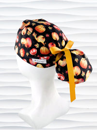 Ponytail style surgical scrub cap hat with orange and yellow pumpkins and leaves tossed on black premium fabric.