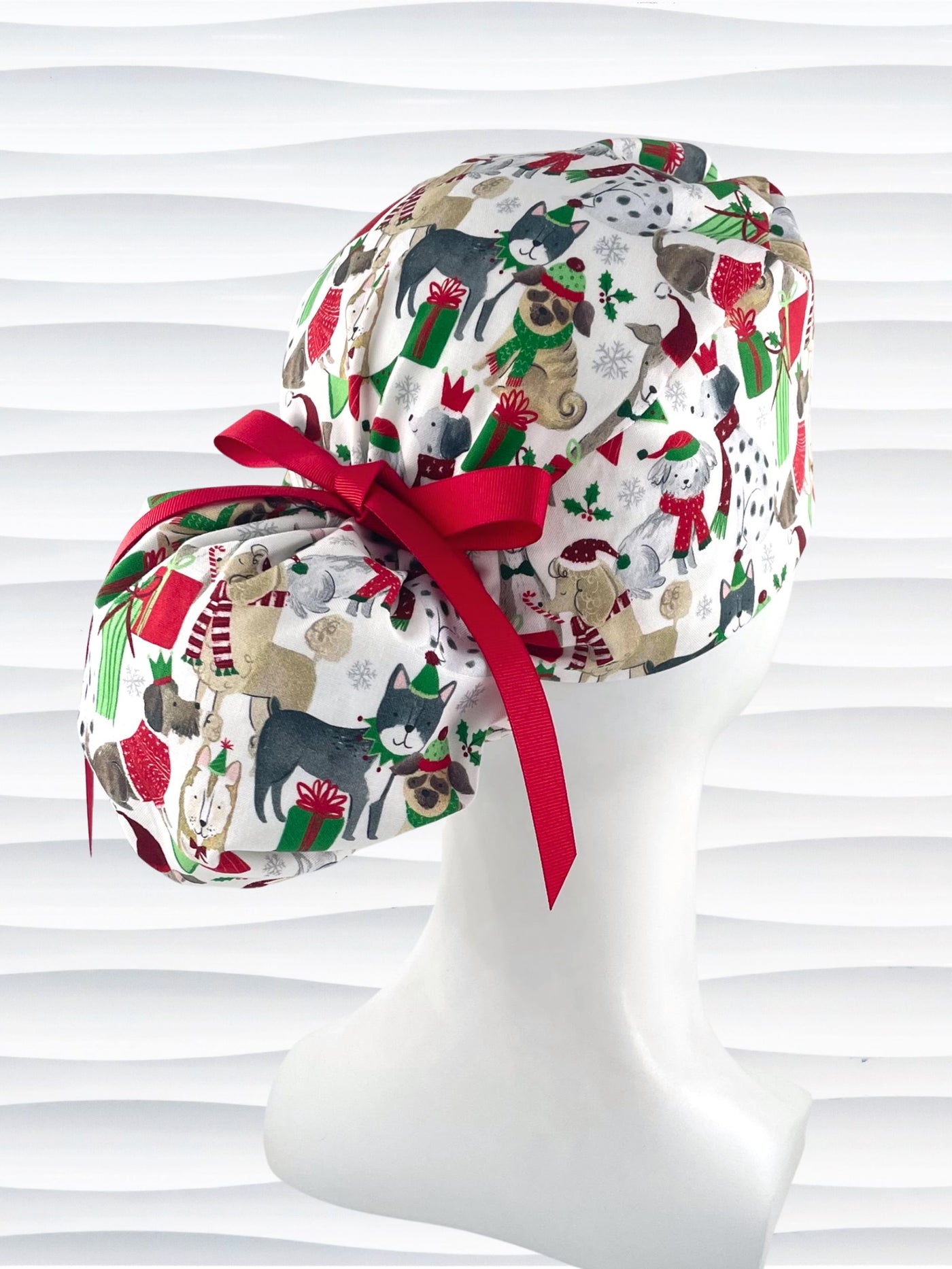 Ponytail style surgical scrub cap hat with dogs in scarves and santa hats in red, green, and white next to snowflakes and christmas presents on white cotton fabric.