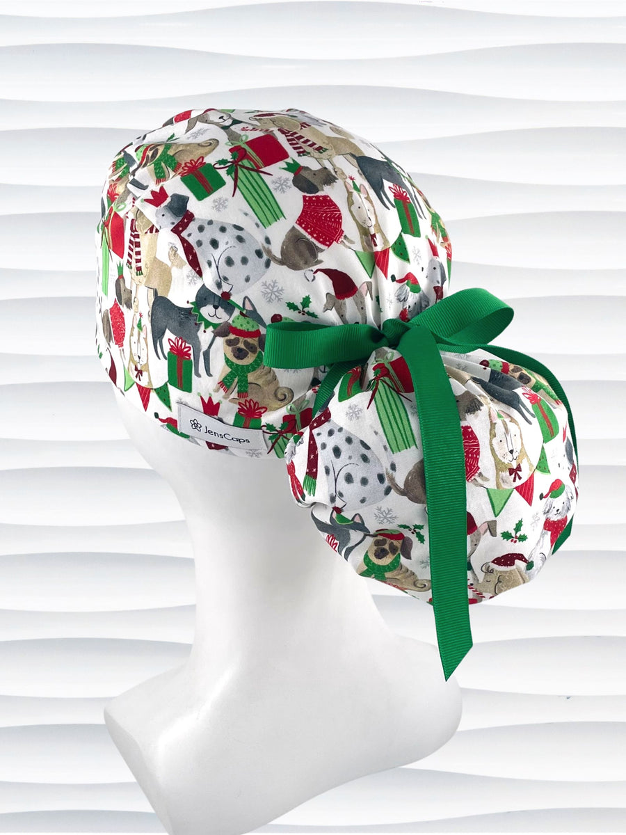 Ponytail style surgical scrub cap hat with dogs in scarves and santa hats in red, green, and white next to snowflakes and christmas presents on white cotton fabric.