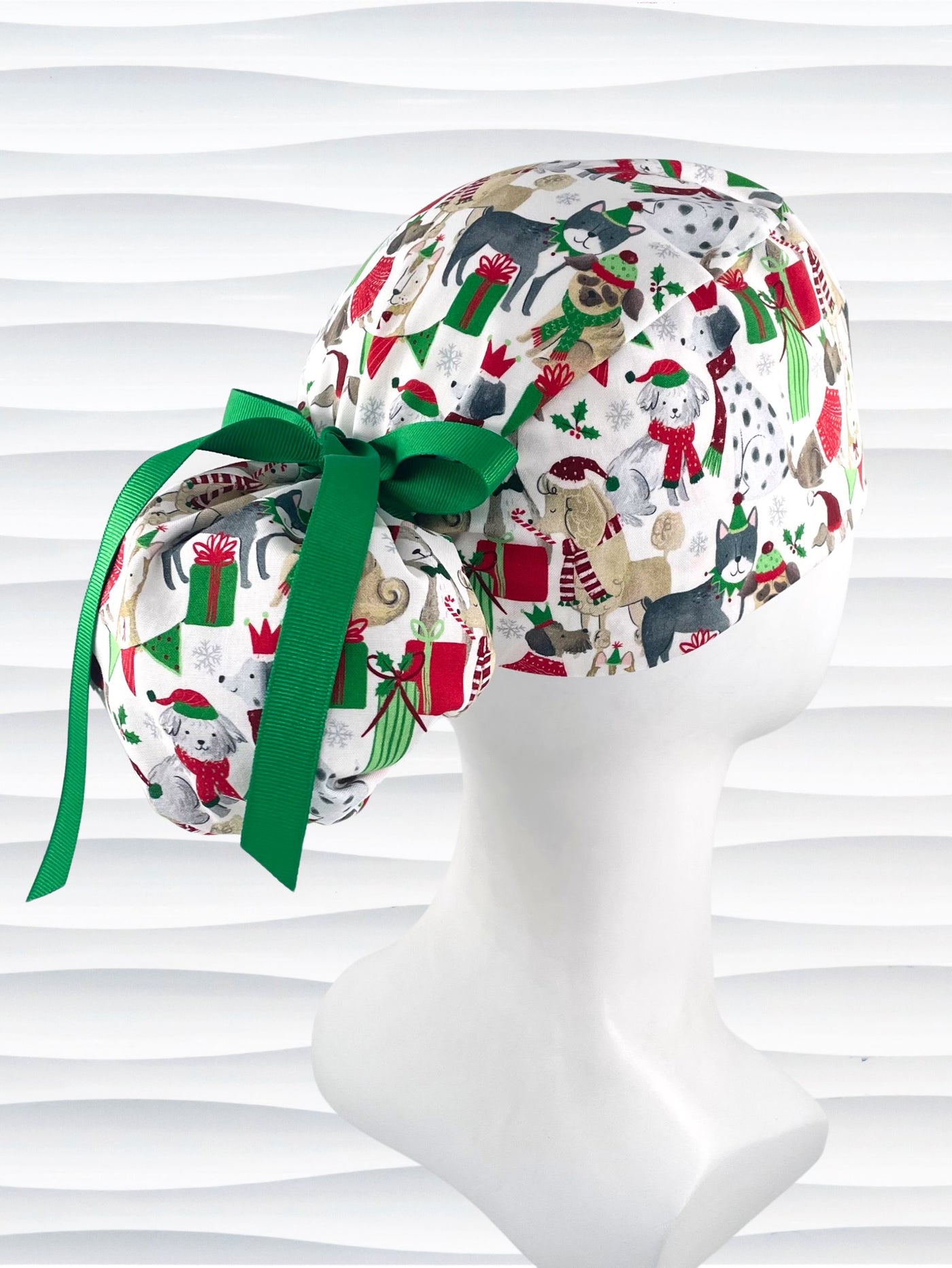 Ponytail style surgical scrub cap hat with dogs in scarves and santa hats in red, green, and white next to snowflakes and christmas presents on white cotton fabric.