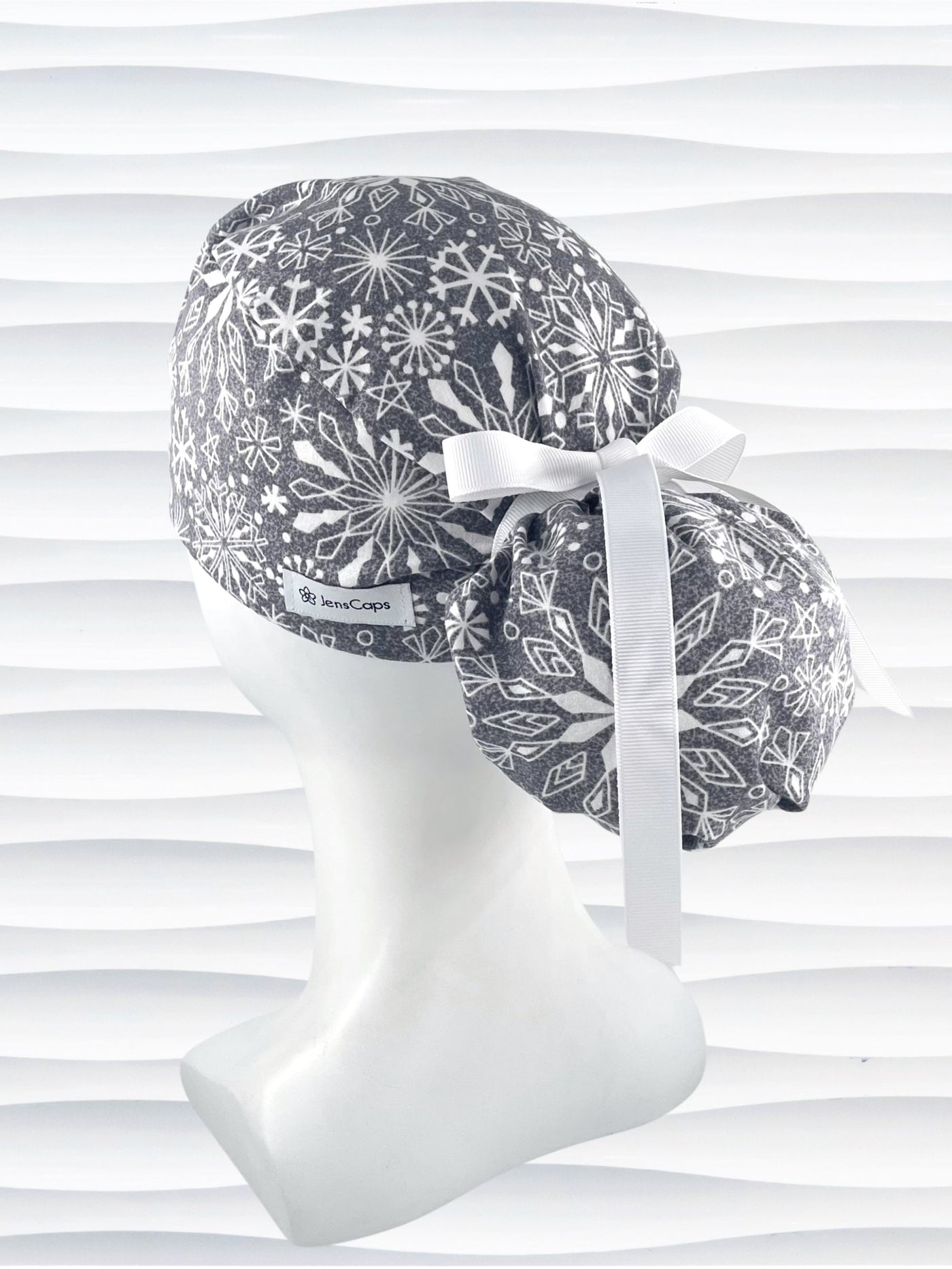 Ponytail style surgical scrub cap hat with white snowflakes all over a light gray cotton fabric.