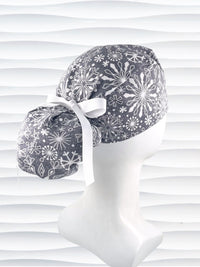 Ponytail style surgical scrub cap hat with white snowflakes all over a light gray cotton fabric.