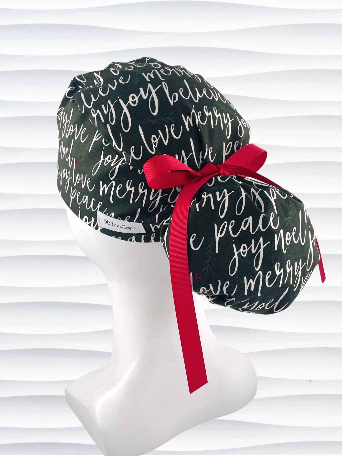 Ponytail style surgical scrub cap hat with love, merry, beleive, joy, peace and noel in a white script on dark green cotton fabric.