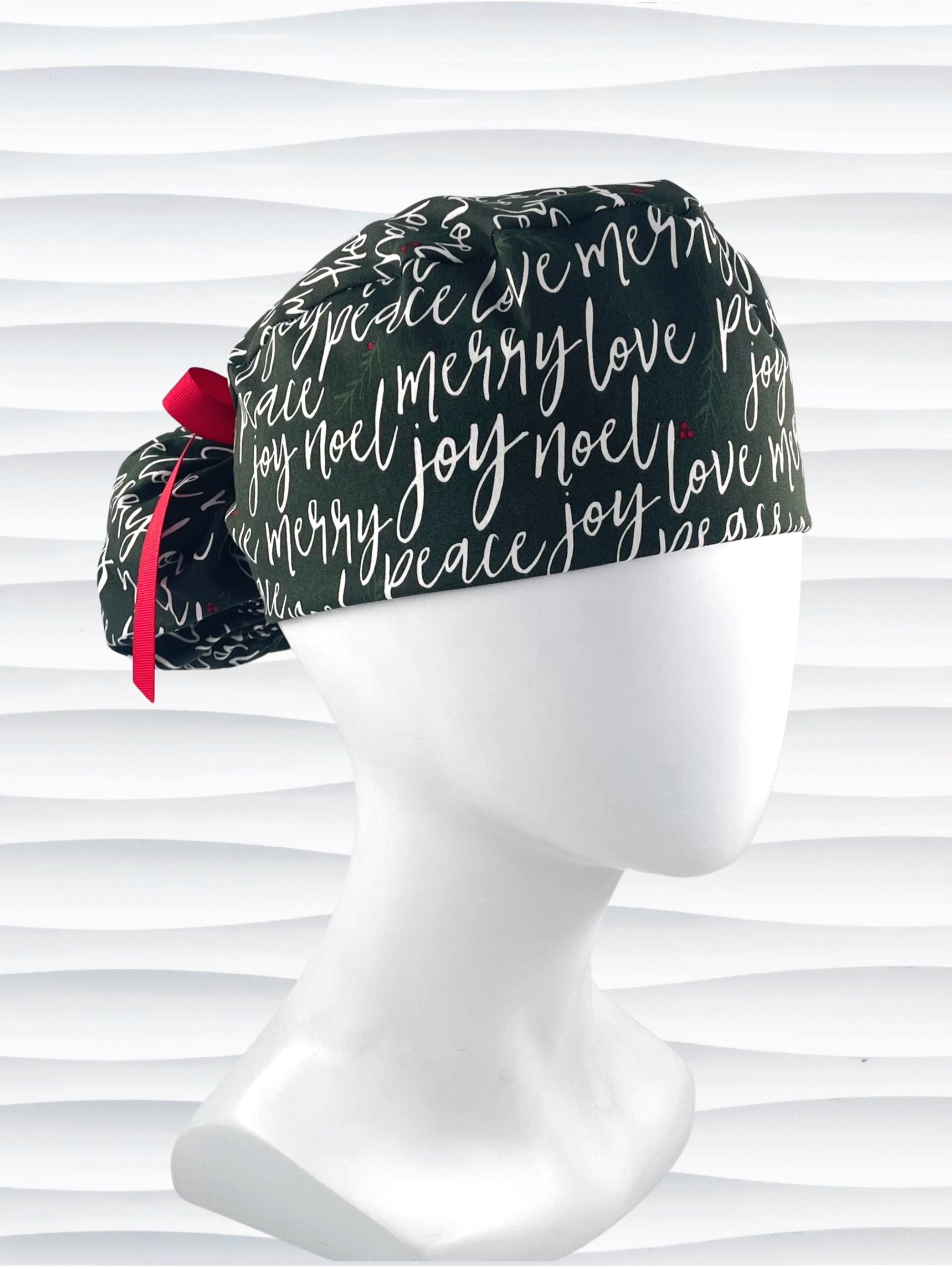 Ponytail style surgical scrub cap hat with love, merry, beleive, joy, peace and noel in a white script on dark green cotton fabric.