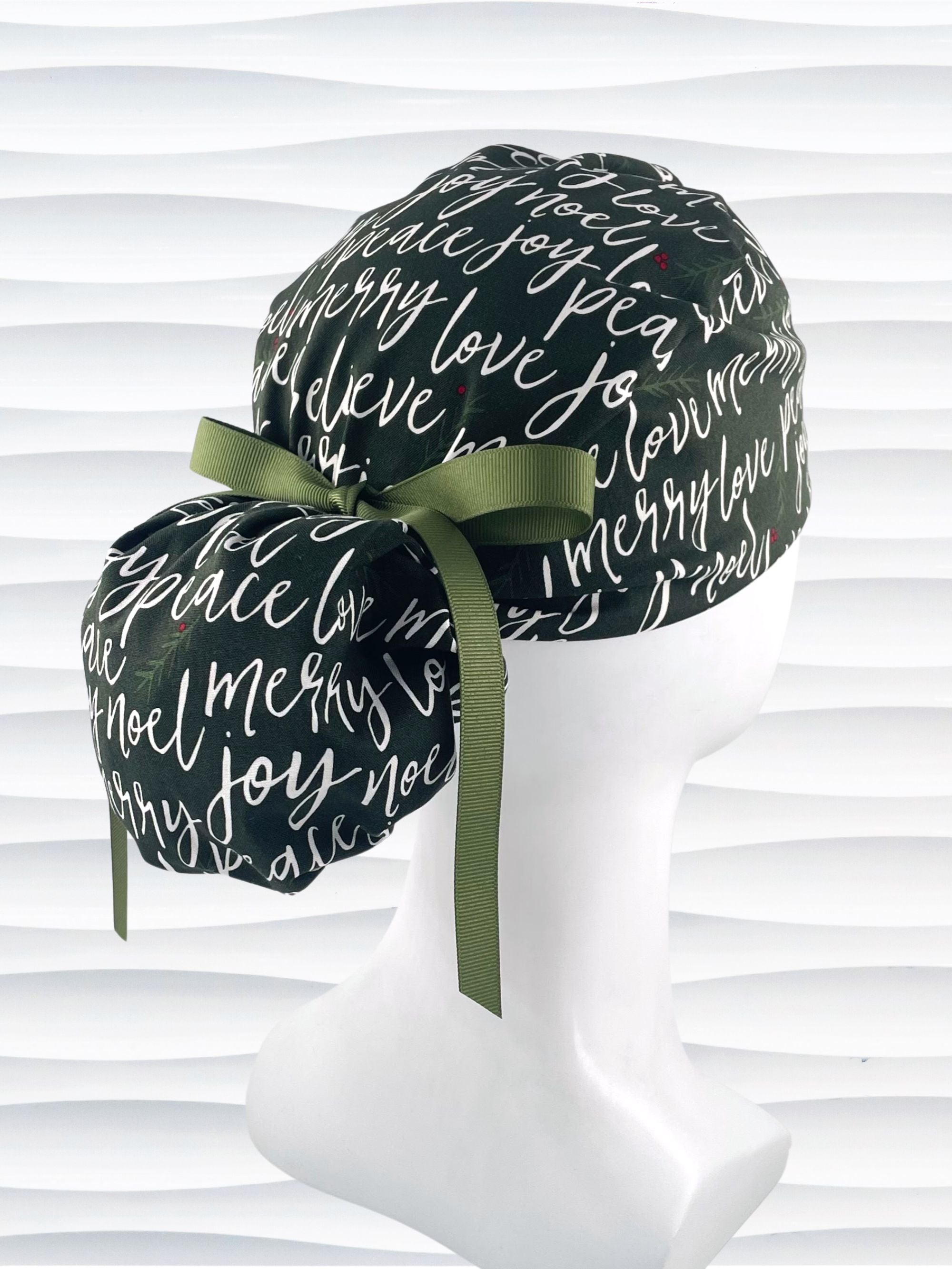 Ponytail style surgical scrub cap hat with love, merry, beleive, joy, peace and noel in a white script on dark green cotton fabric.